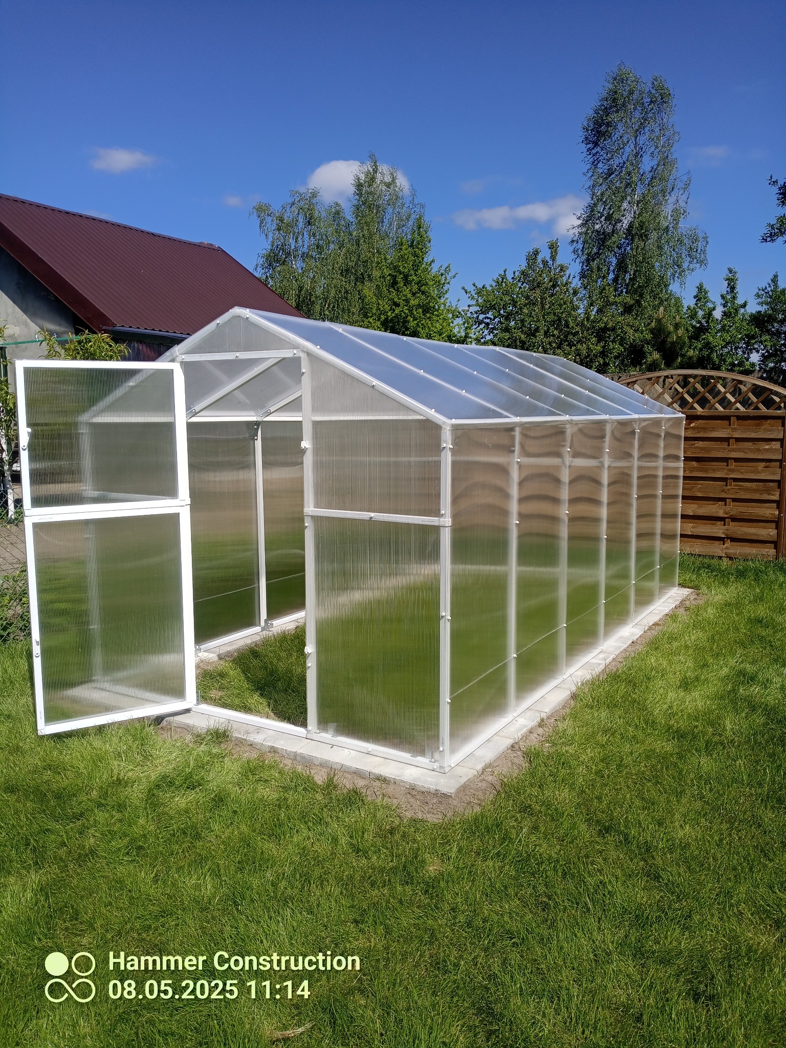 Nowoczesna szklarnia z poliwęglanu na trawniku, z otwartymi drzwiami, widok na błękitne niebo i dom w tle. Konstrukcja z białych profili.