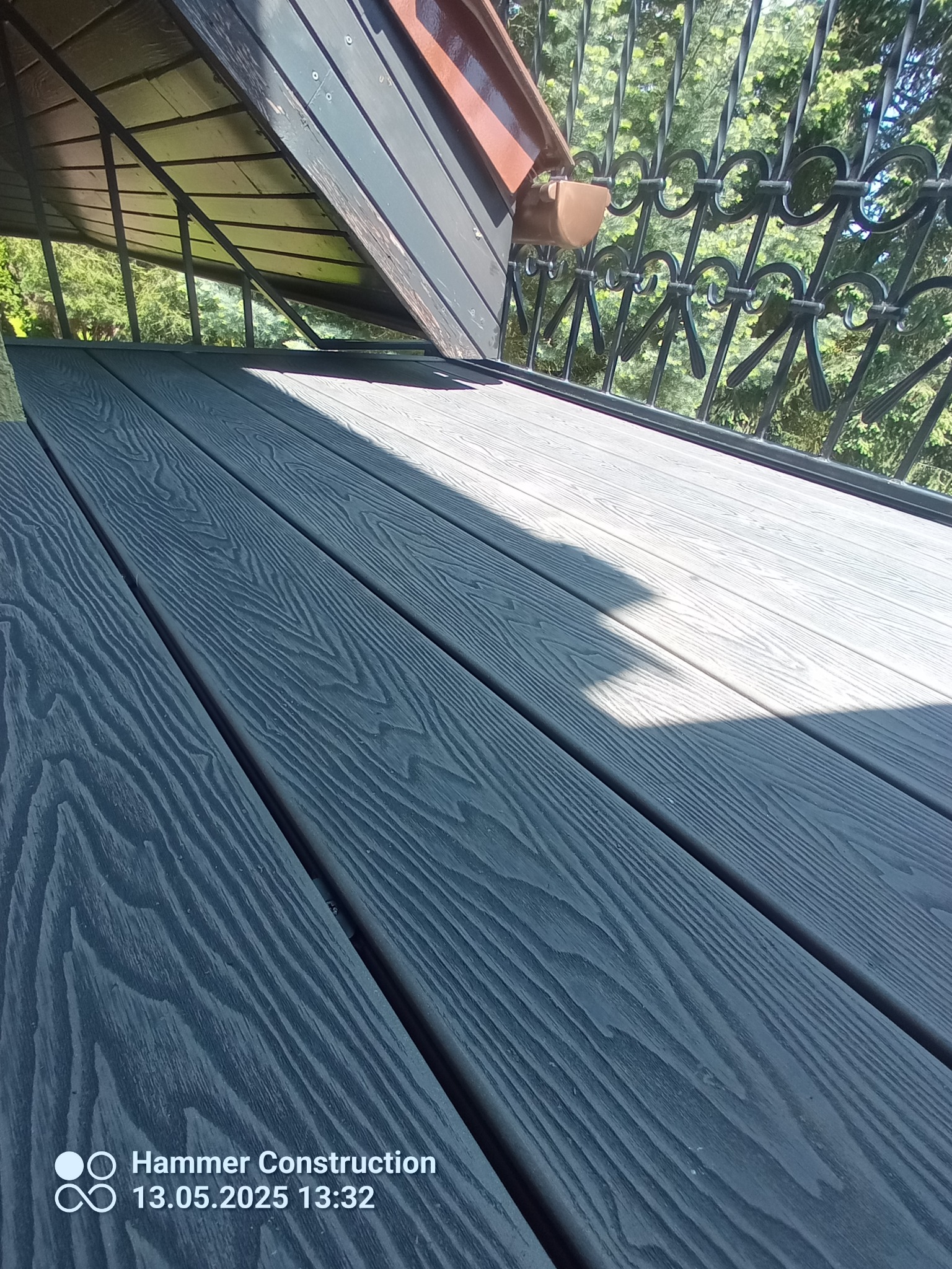 Szare deski kompozytowe na tarasie z metalową balustradą w tle, widok z góry, oświetlone słońcem, z widoczną fakturą drewna i cieniem.