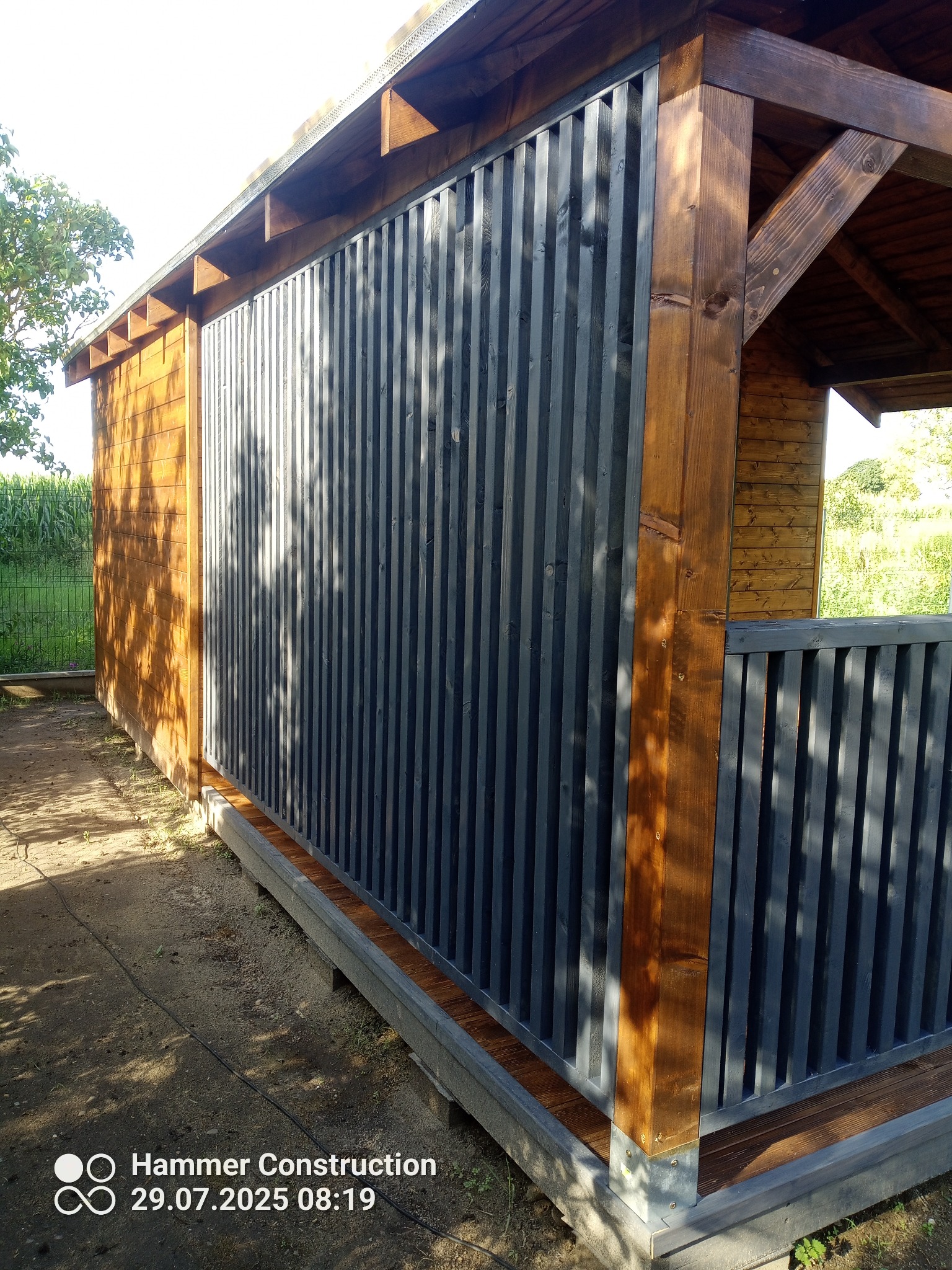 Drewniana konstrukcja z szarymi, pionowymi panelami, prawdopodobnie altana lub sauna, z widocznymi elementami drewnianymi i betonową podstawą. Ujęcie z zewnątrz.