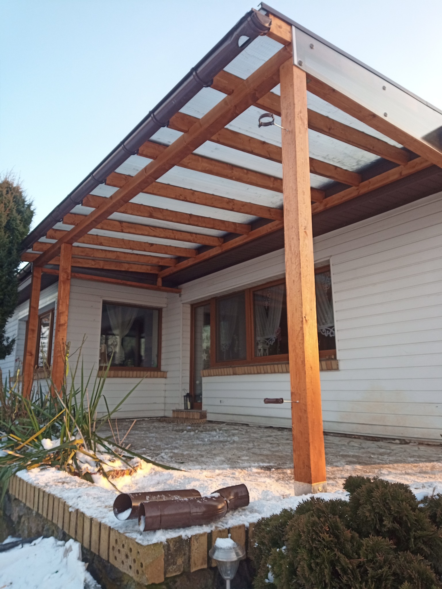 Drewniana pergola z przezroczystym zadaszeniem przy białym domu. Widoczne elementy konstrukcyjne, rynna i fragment ogrodu z resztkami śniegu. Ujęcie w słoneczny dzień.