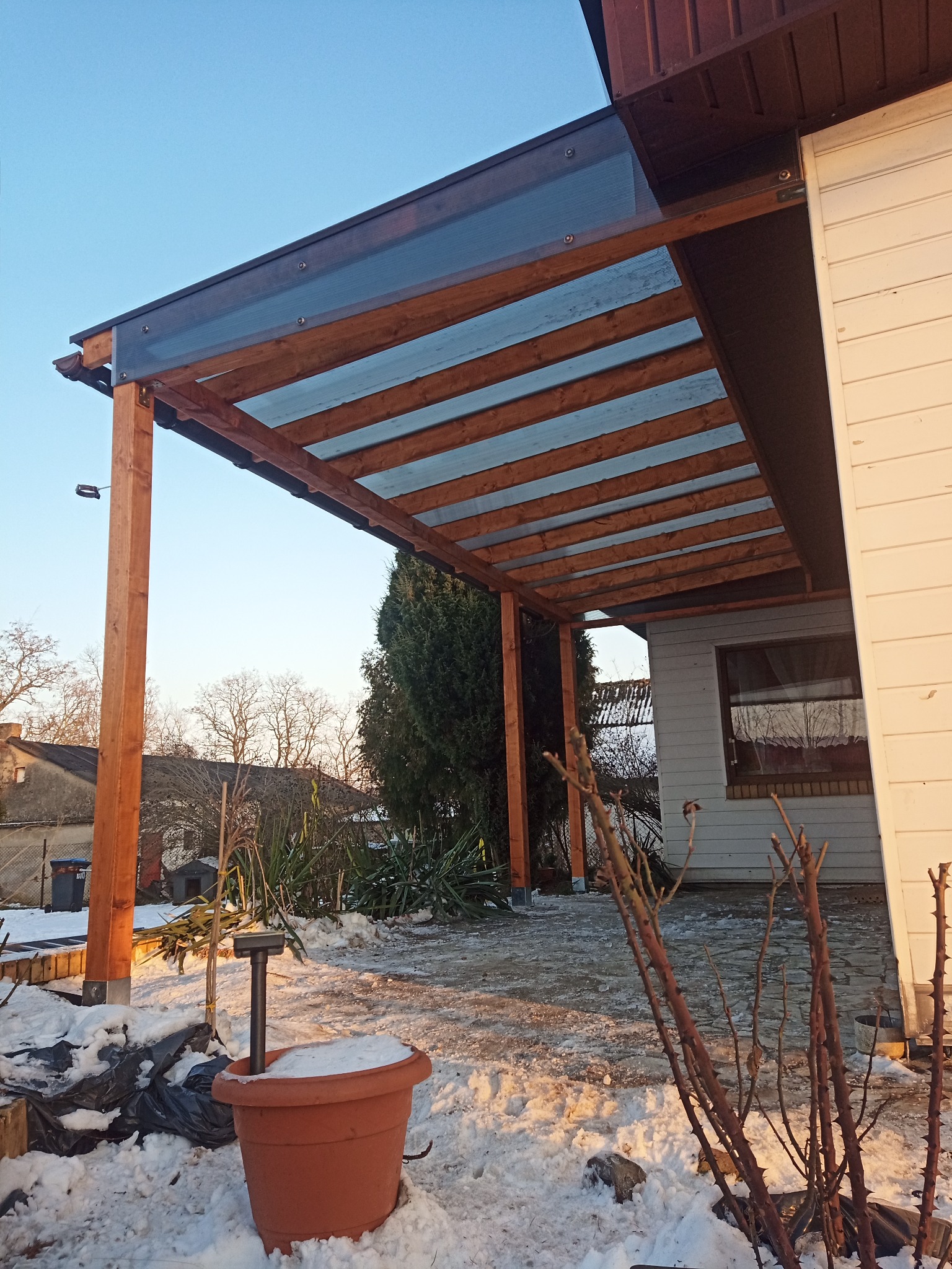 Drewniana pergola z przezroczystym zadaszeniem, widok z dołu, na pierwszym planie donica z pokrywą śnieżną, zimowy krajobraz w tle, błękitne niebo.