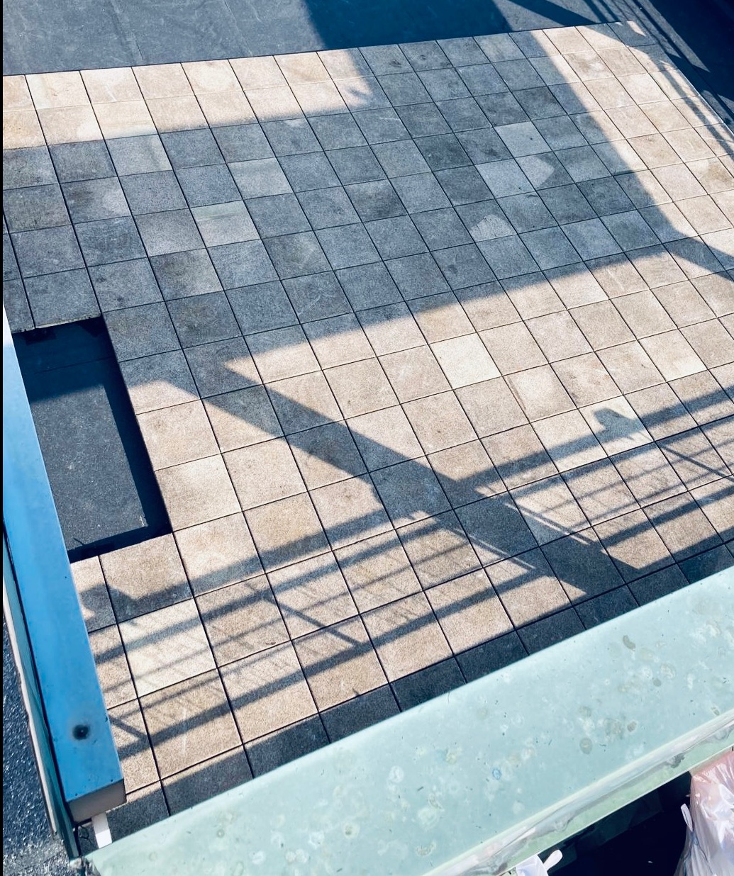 Szare płyty dachowe ułożone w kwadraty, z brakującą płytą w rogu i cieniami rzucanymi przez barierki, widok z góry.