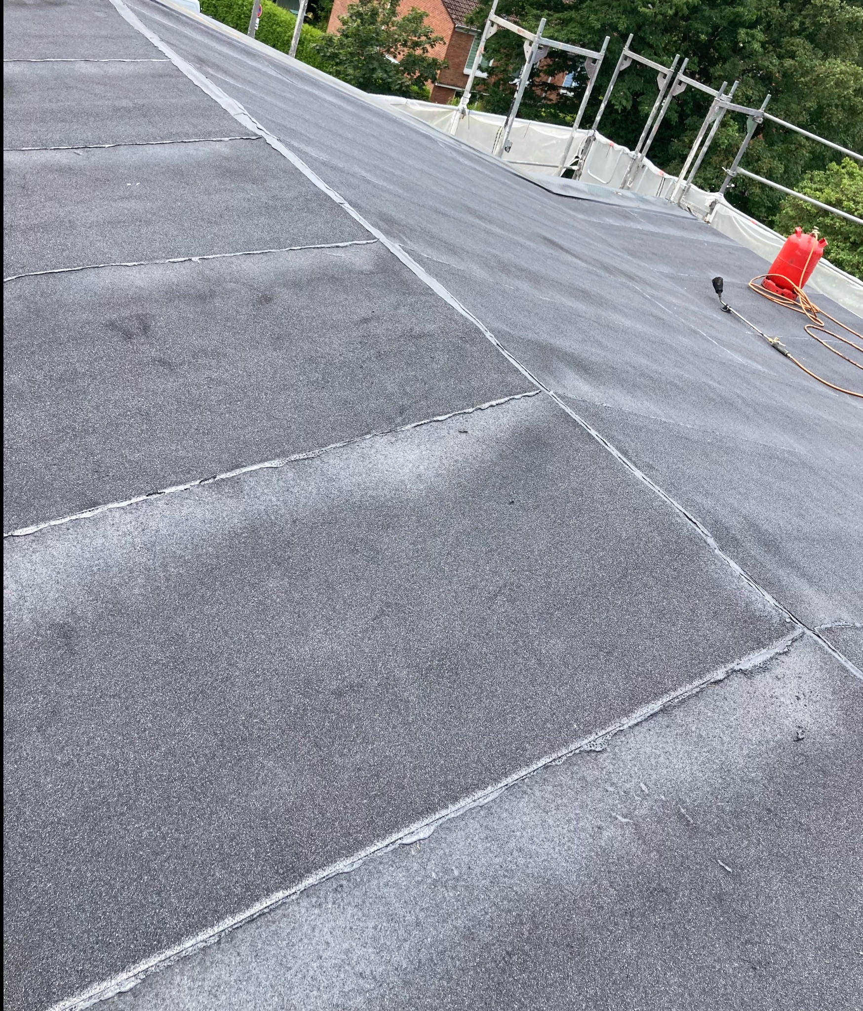 Czarny, płaski dach pokryty papą termozgrzewalną, widoczne łączenia, w tle rusztowanie i zieleń.