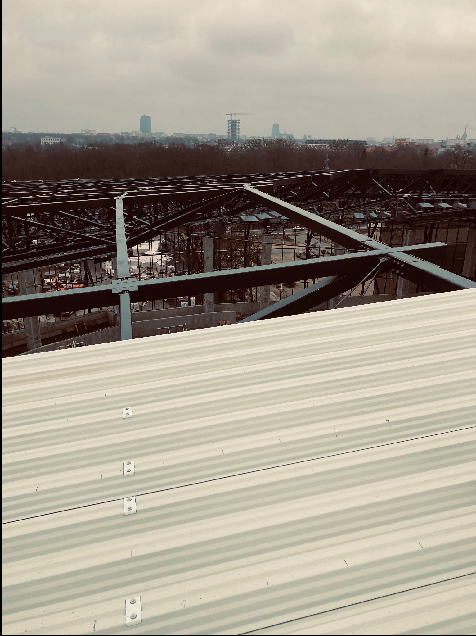 Widok z góry na nową, jasną blachę falistą na dachu w trakcie budowy, z konstrukcją stalową i panoramą miasta w tle, pochmurny dzień.