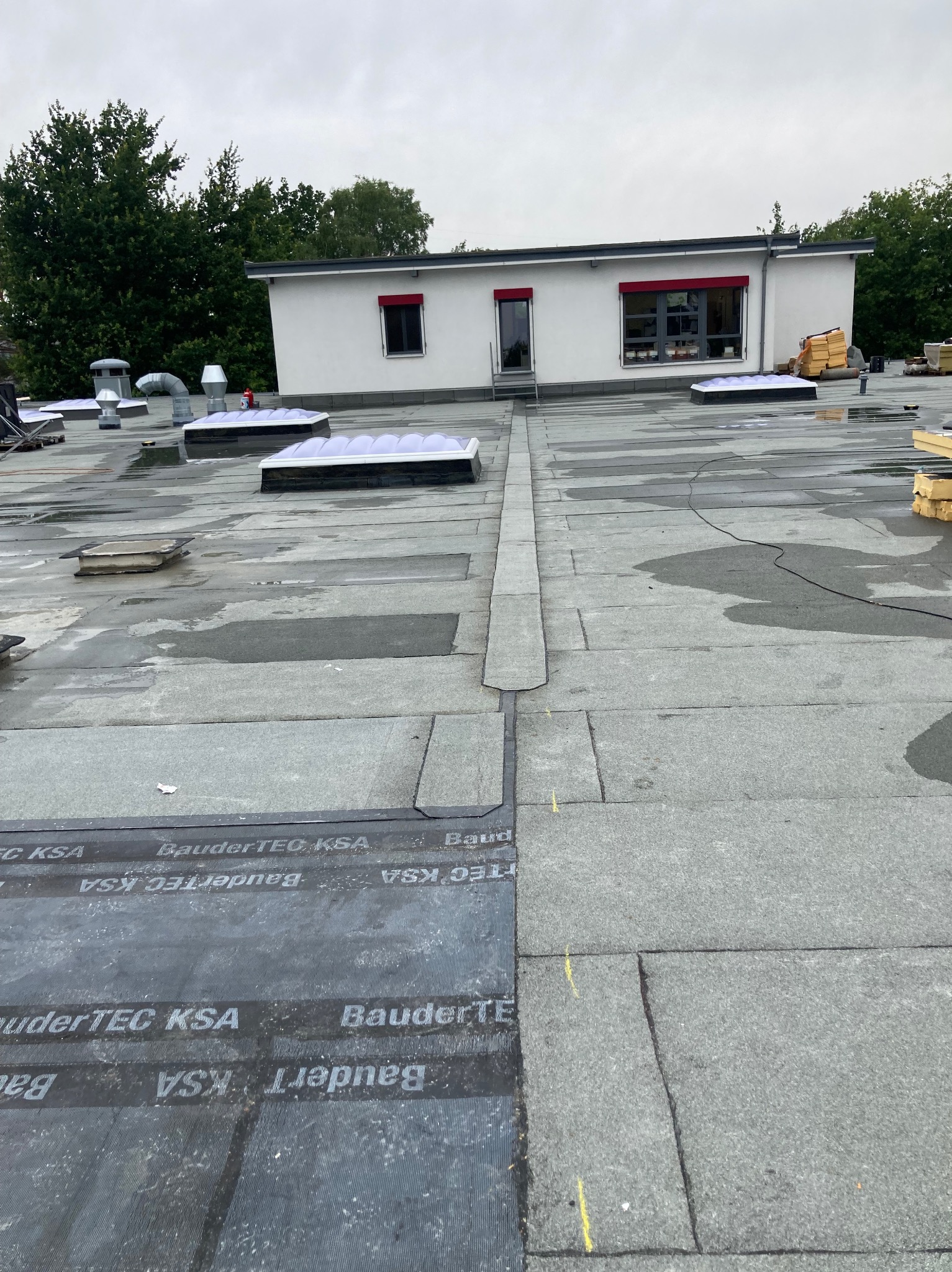 Płaski dach pokryty papą termozgrzewalną z widocznymi świetlikami i elementami wentylacji. Fragment budynku w tle. Widoczne łączenia papy i detale wykończeniowe.