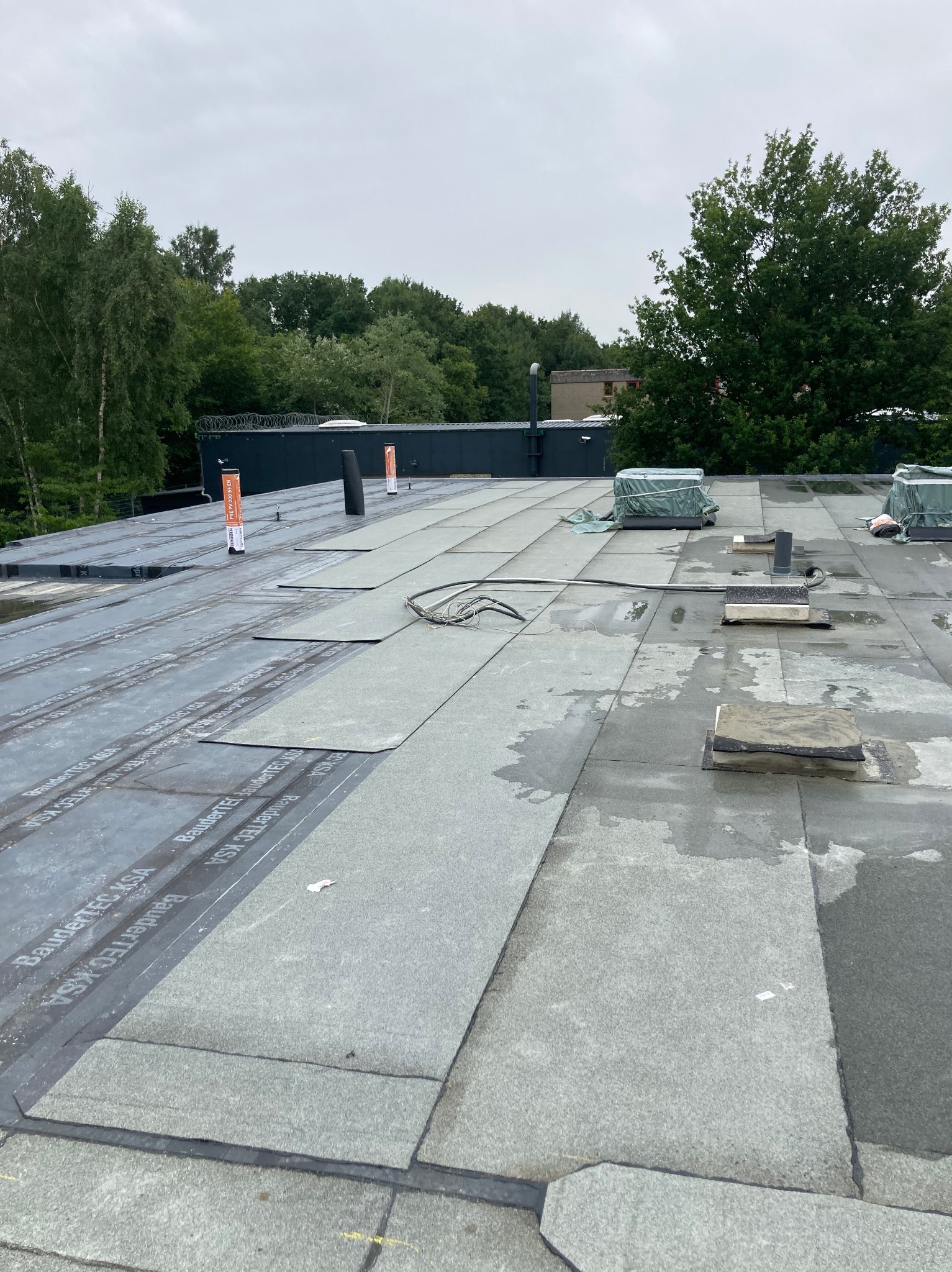 Płaski dach pokryty szarą papą z widocznymi łączeniami, elementami wentylacyjnymi i przewodami. W tle zieleń drzew i fragment budynku z drutem kolczastym.