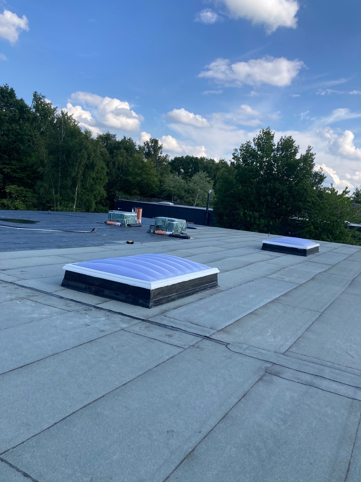 Płaski dach pokryty papą z trzema świetlikami dachowymi, widok z góry na tle zieleni i błękitnego nieba z chmurami.