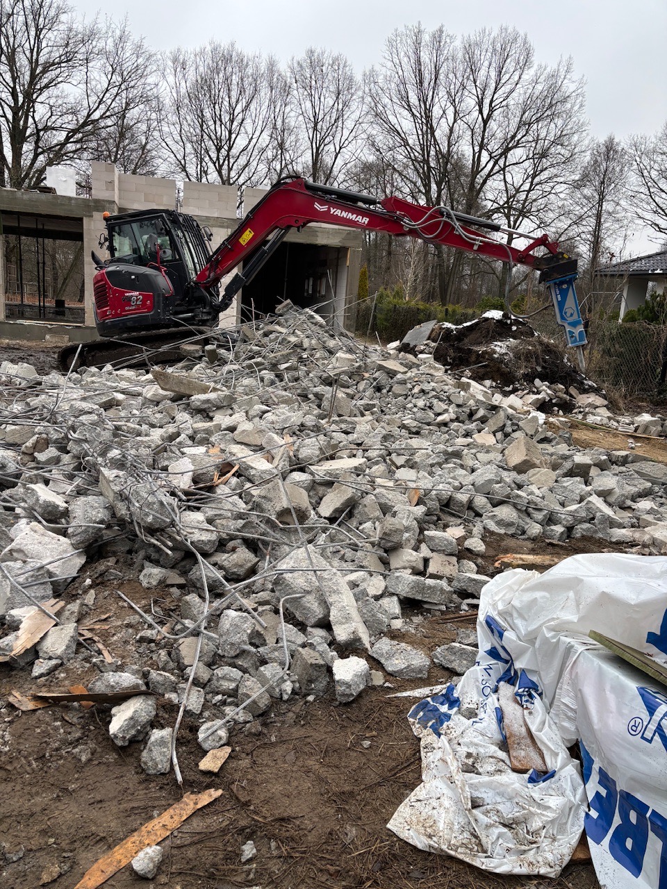 Czerwony minikoparka Yanmar z młotem pneumatycznym rozbija betonowe elementy na placu budowy. W tle niedokończony budynek i drzewa bez liści.