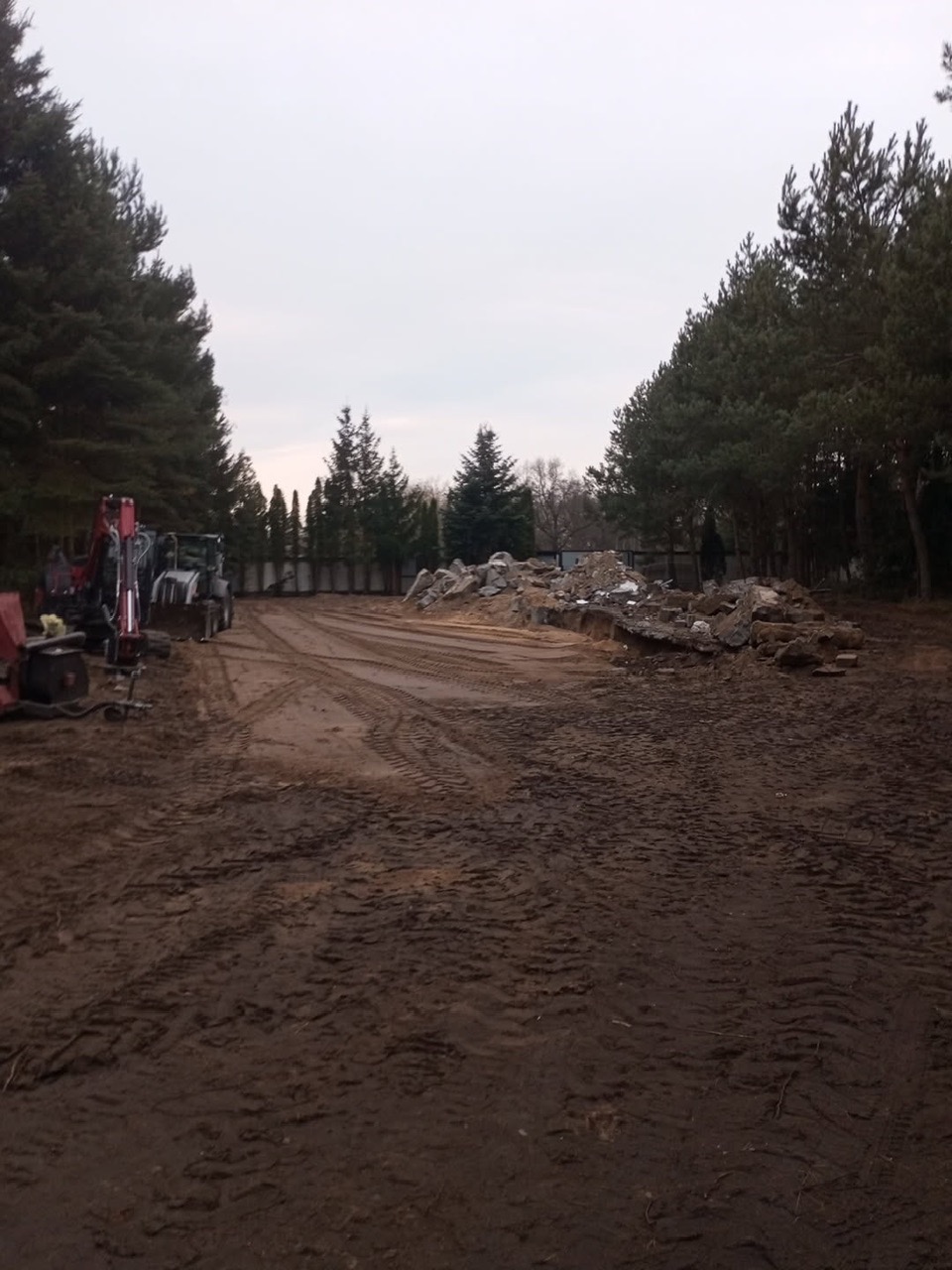 Teren po rozbiórce z gruzem i śladami opon, widoczne maszyny budowlane i drzewa w tle, pochmurne niebo. Etap prac przygotowawczych pod budowę.