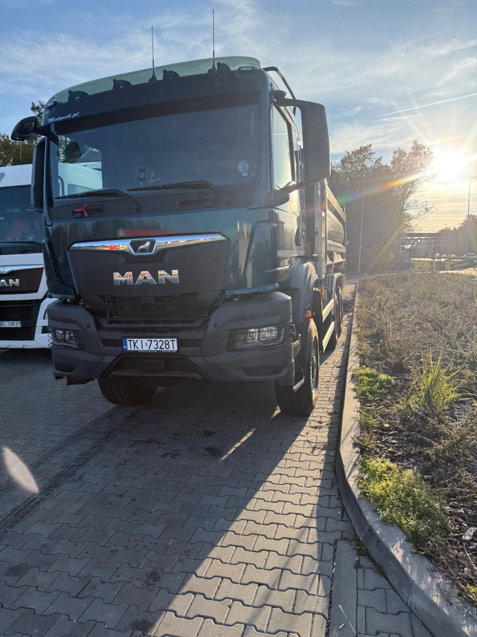 Ciężarówka MAN w zielonym kolorze, zaparkowana na kostce brukowej w słoneczny dzień. Widoczna tablica rejestracyjna TKI 7328T i logo MAN. Częściowo widoczna inna ciężarówka.