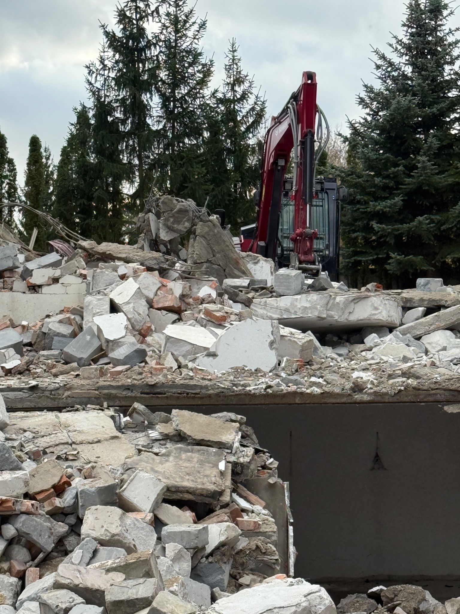 Częściowo zburzony budynek z gruzem i czerwonym ramieniem koparki na tle drzew iglastych pod pochmurnym niebem. Widoczne fragmenty ścian i stropu.