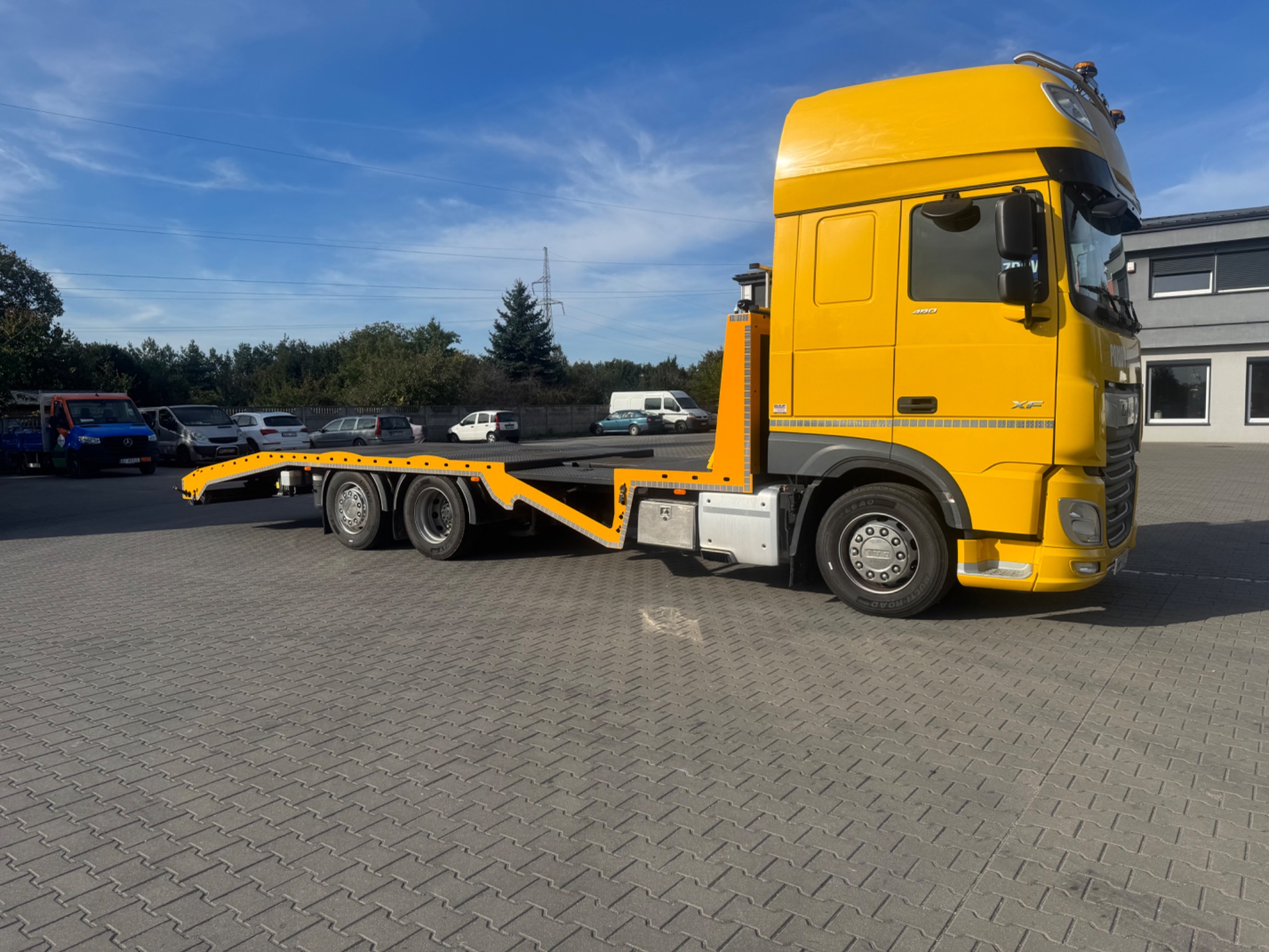 Żółta laweta DAF XF na szarym bruku, widok z boku, błękitne niebo w tle. Pojazdy osobowe zaparkowane w oddali. Charakterystyczny, jaskrawy kolor lawety.