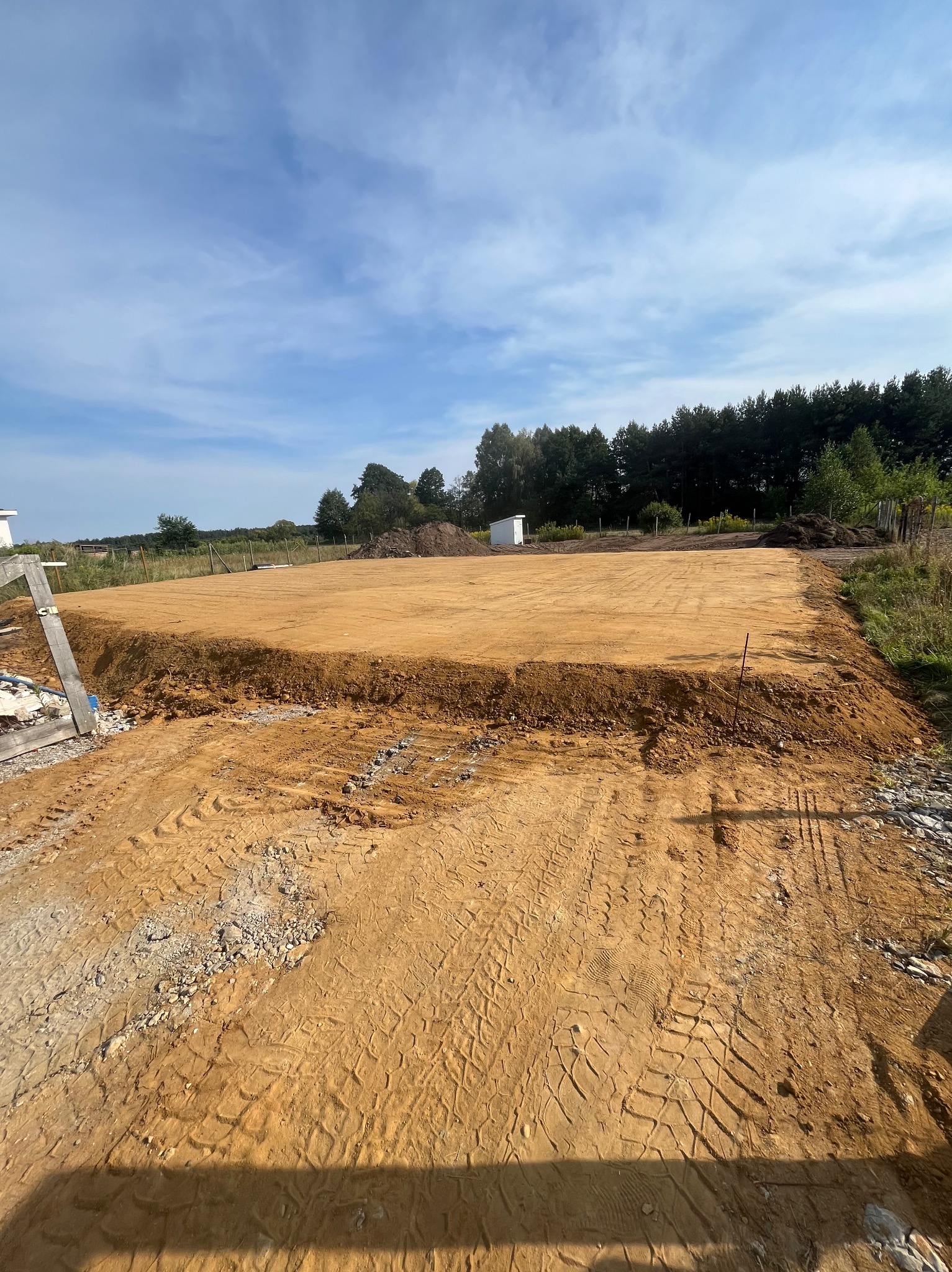 Przygotowany teren pod budowę z widocznymi śladami opon, wyrównana powierzchnia ziemi, w tle drzewa i błękitne niebo z chmurami. Widoczne narzędzia i materiały budowlane.