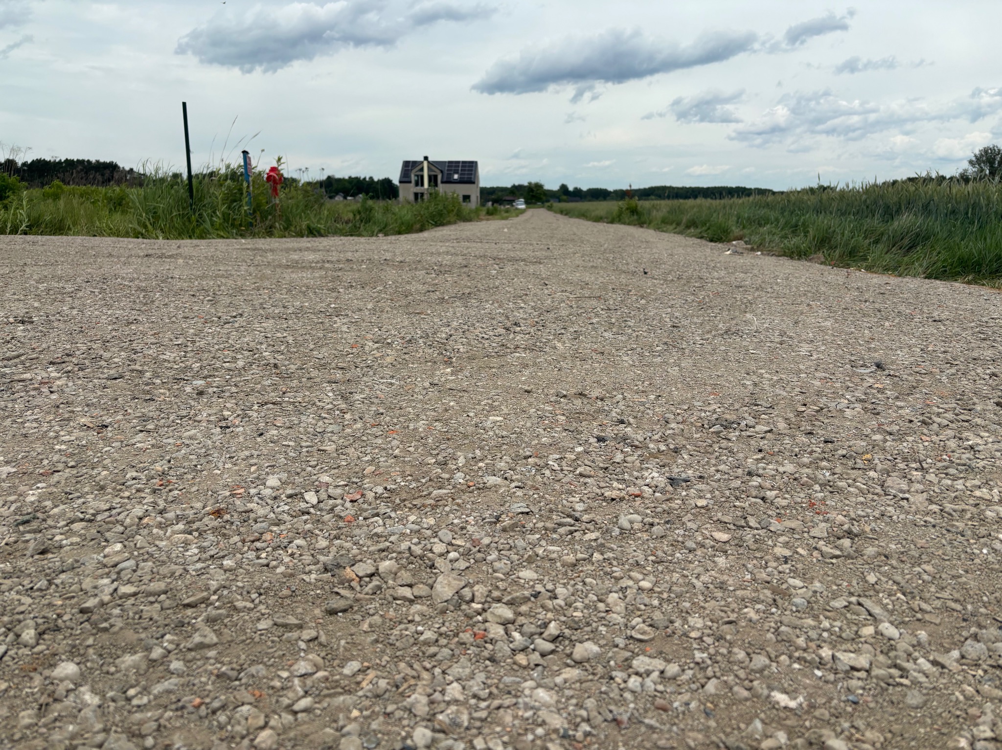 Szutrowa droga dojazdowa z widocznym domem w tle, otoczona zielenią. Perspektywa z poziomu gruntu podkreśla fakturę nawierzchni i rozległość terenu.