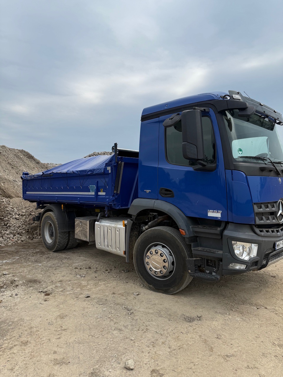 Niebieski samochód ciężarowy Mercedes-Benz z plandeką, zaparkowany na placu budowy z widoczną stertą gruzu w tle, pochmurne niebo.