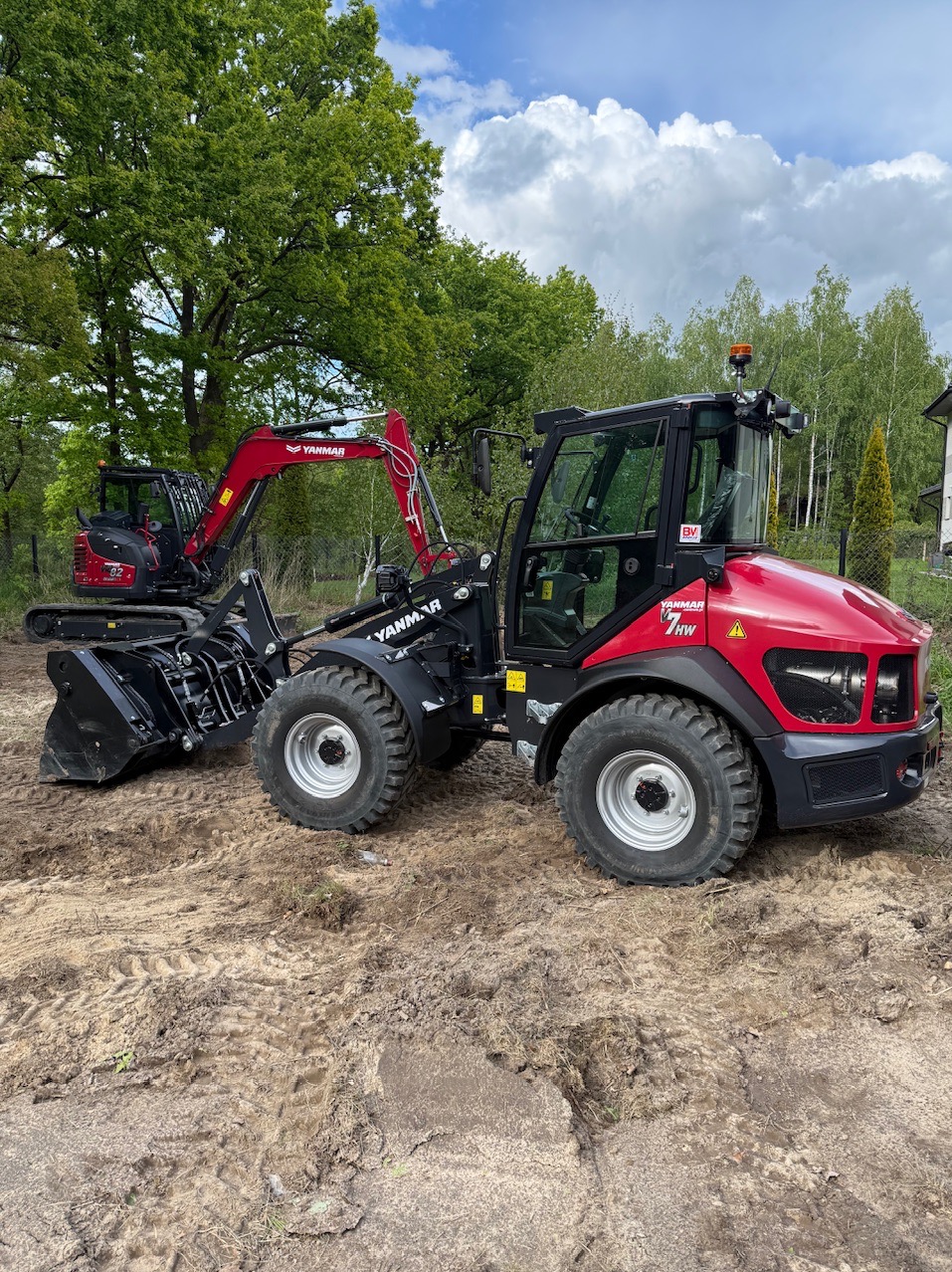 Czerwony ciągnik Yanmar z łyżką i koparka na ziemi, otoczone zielenią. Sprzęt budowlany gotowy do pracy, z widocznymi śladami opon na gruncie.
