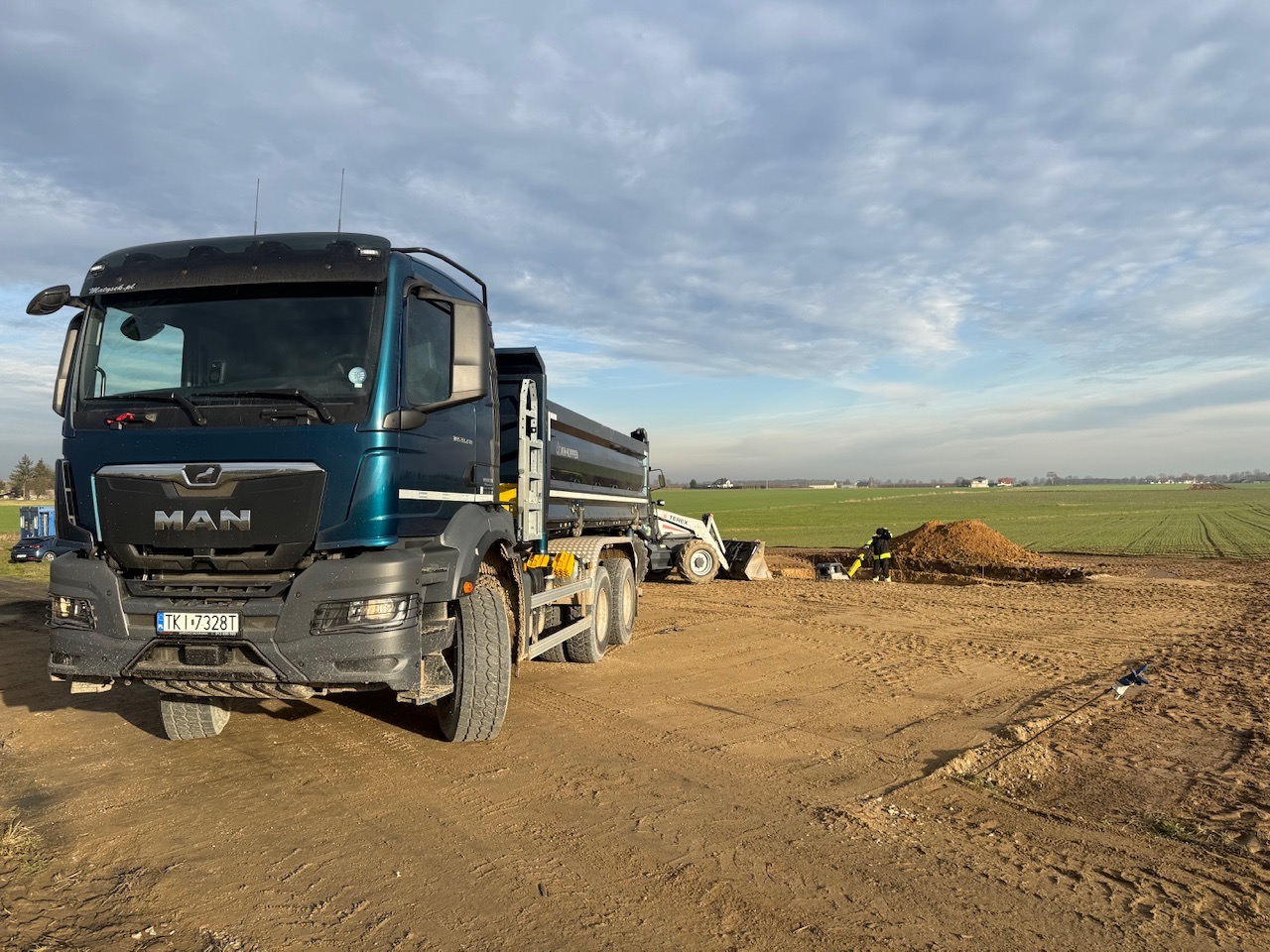Ciężarówka MAN wywrotka na placu budowy, w tle prace ziemne z użyciem małej ładowarki i pracownika w kombinezonie. Krajobraz polny pod pochmurnym niebem.
