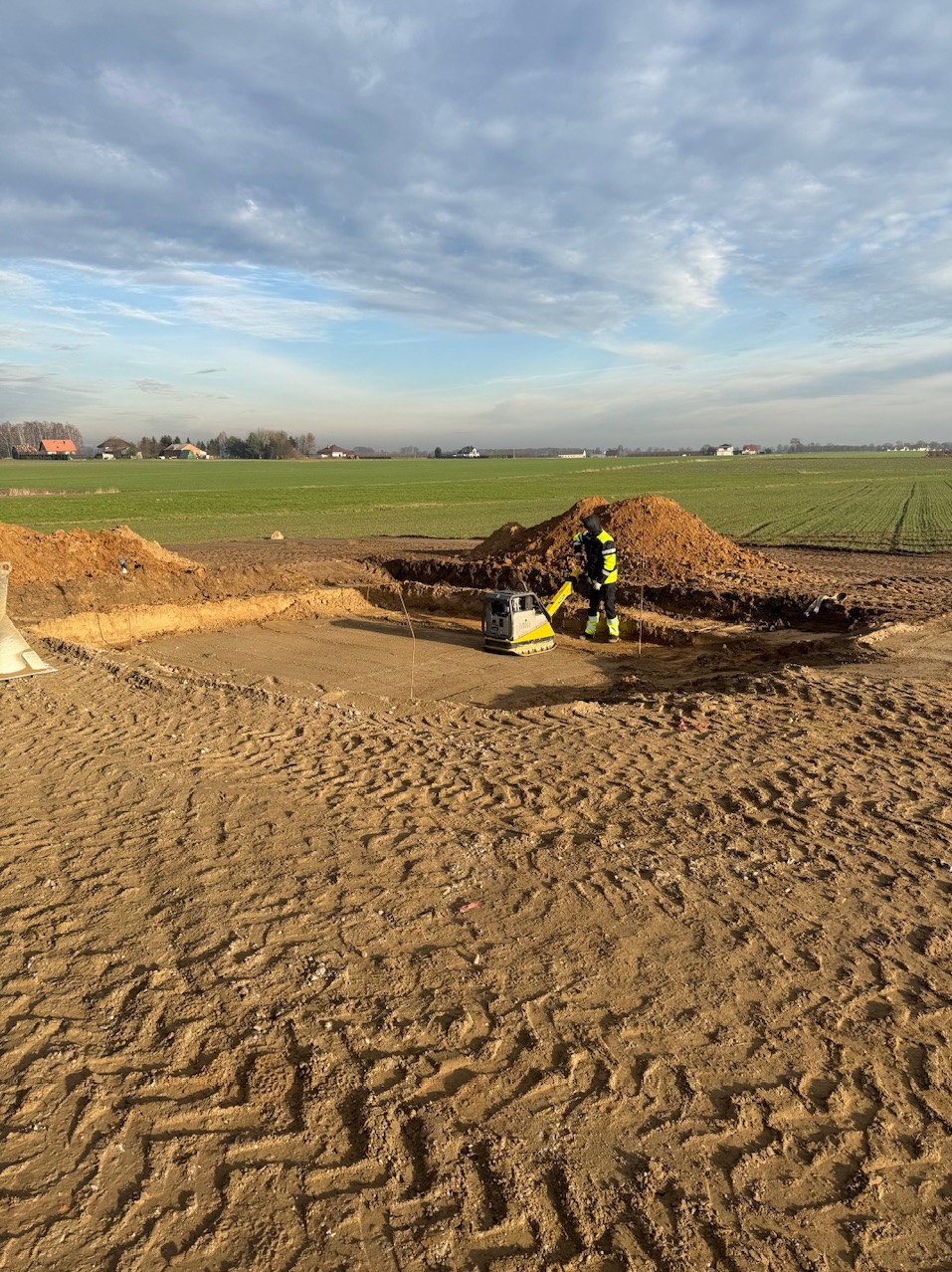 Prace ziemne w Wyszkowie: zagęszczarka w akcji przy wykopie pod fundamenty, widoczne ślady opon i hałdy ziemi na tle krajobrazu.