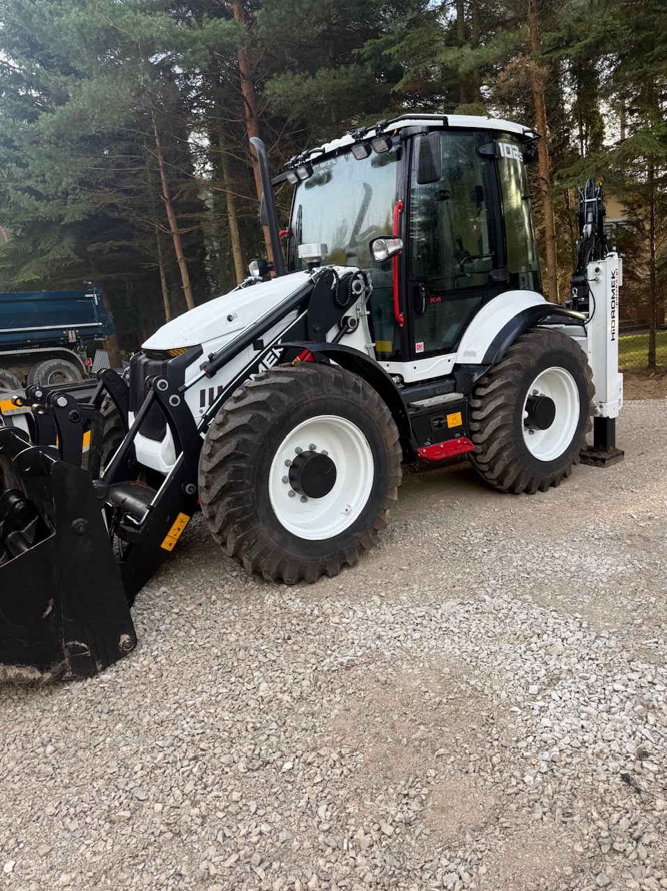 Nowoczesna, biało-czarna koparko-ładowarka Hidromek na żwirowym podłożu, z widocznym łyżką i wysięgnikiem, na tle drzew. Sprzęt gotowy do pracy.