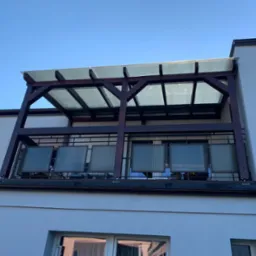 Zadaszony balkon z drewnianą konstrukcją i przezroczystym dachem, widok z dołu na tle błękitnego nieba.