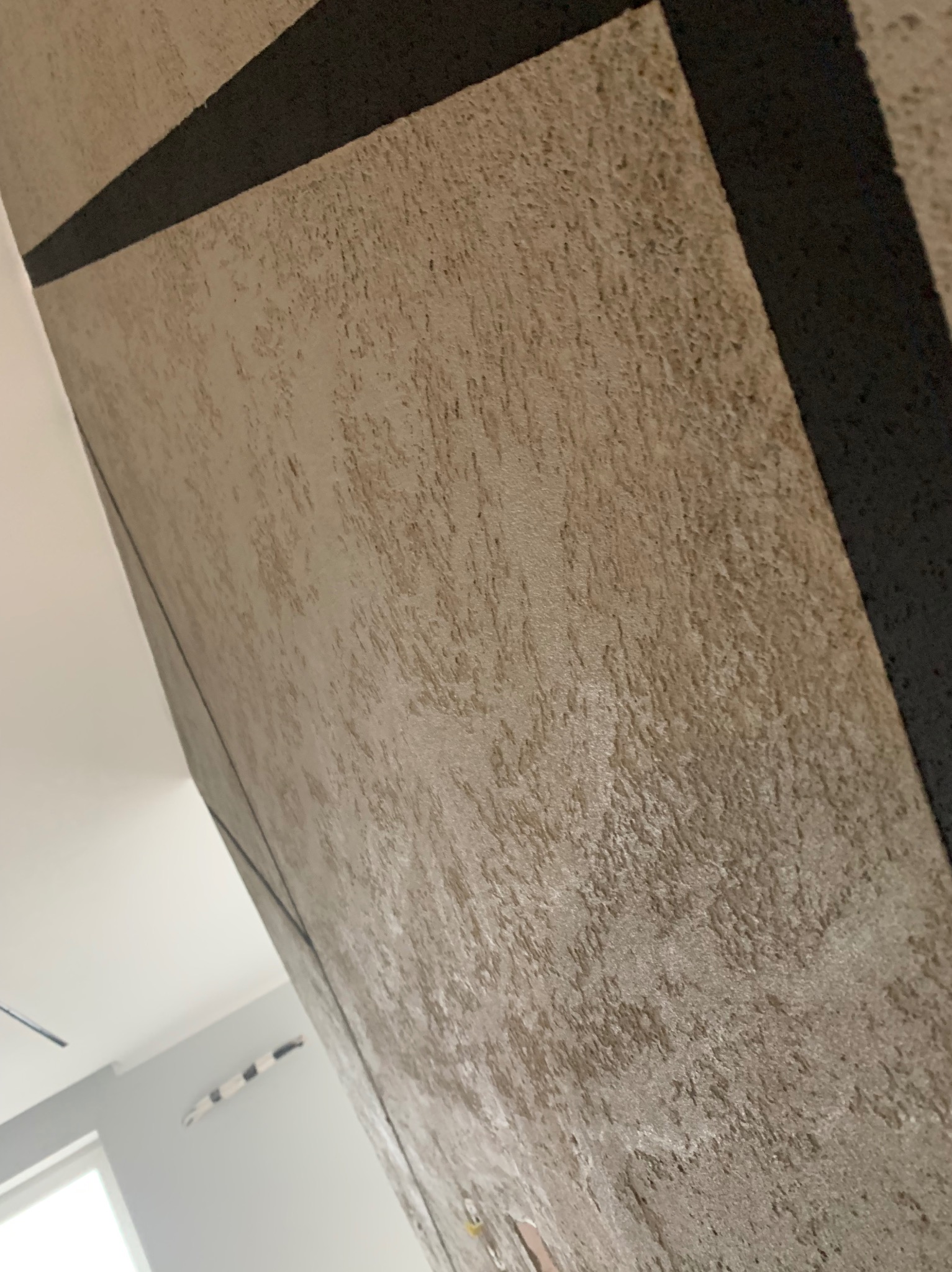 Szara ściana z dekoracyjną strukturą i czarnym pasem, widoczna z bliska pod kątem, fragment sufitu i okna w tle.