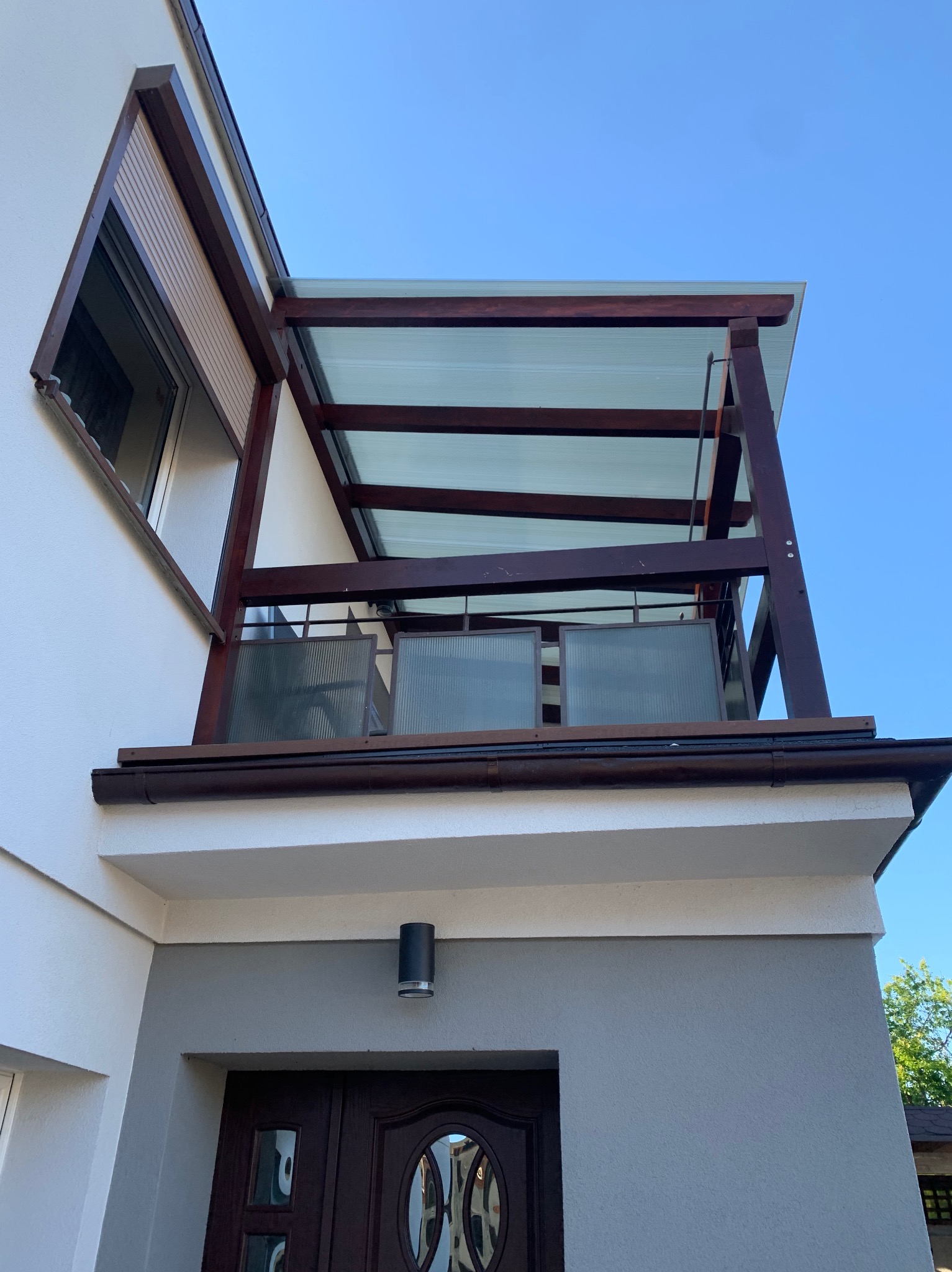 Zadaszenie balkonu z brązowymi elementami drewnianymi i przezroczystym dachem, widok z dołu na tle błękitnego nieba.