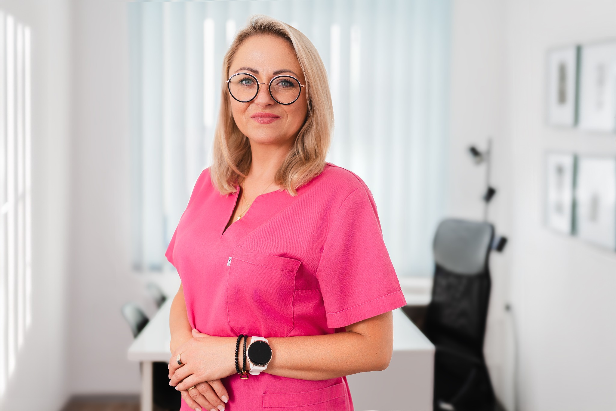 Portret uśmiechniętej kobiety w różowym uniformie medycznym i okularach, stojącej na tle jasnego biura z biurkiem i krzesłem.