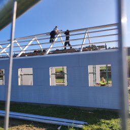 PHU KAROLINA SOKALSKA - Montaż konstrukcji stalowej dachu na budynku w trakcie budowy, widoczne panele ścienne i pracownicy na górze.