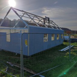 PHU KAROLINA SOKALSKA - Wznoszenie szkieletu domu z prefabrykatów na tle zachodzącego słońca, widoczni dwaj pracownicy na dachu, ogrodzenie budowy z siatki.