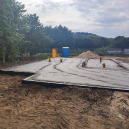 PHU KAROLINA SOKALSKA - Wylana betonowa płyta fundamentowa z wystającymi rurami i zbrojeniami, widoczna izolacja pionowa, w tle przenośna toaleta i pryzma piasku na placu budowy.