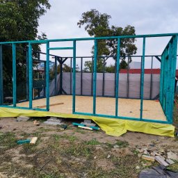 PHU KAROLINA SOKALSKA - Wznoszenie szkieletu domu: widok na pomalowaną na turkusowo drewnianą konstrukcję ścian z płytami OSB na podłodze, zabezpieczoną żółtą folią budowlaną, na tle zieleni i pochmurnego nieba.