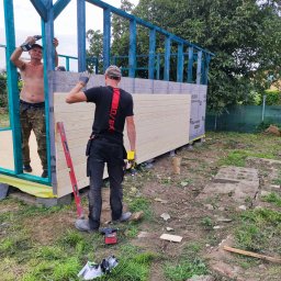 PHU KAROLINA SOKALSKA - Dwóch mężczyzn montuje drewnianą konstrukcję szkieletową z niebieskimi elementami, jeden przykręca śruby od wewnątrz, drugi przykłada poziomicę od zewnątrz, widoczne narzędzia i materiały budowlane.