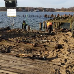 PHU KAROLINA SOKALSKA - Demontaż starego drewnianego pomostu nad jeziorem, pracownicy w kamizelkach odblaskowych podczas pracy, widoczne resztki desek i konstrukcji, w tle woda i drzewa.
