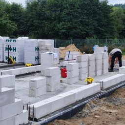 PHU KAROLINA SOKALSKA - Budowa fundamentów z bloczków betonowych komórkowych na placu budowy, widoczny pracownik przygotowujący zaprawę, palety z bloczkami, izolacja fundamentów i zbrojenie.