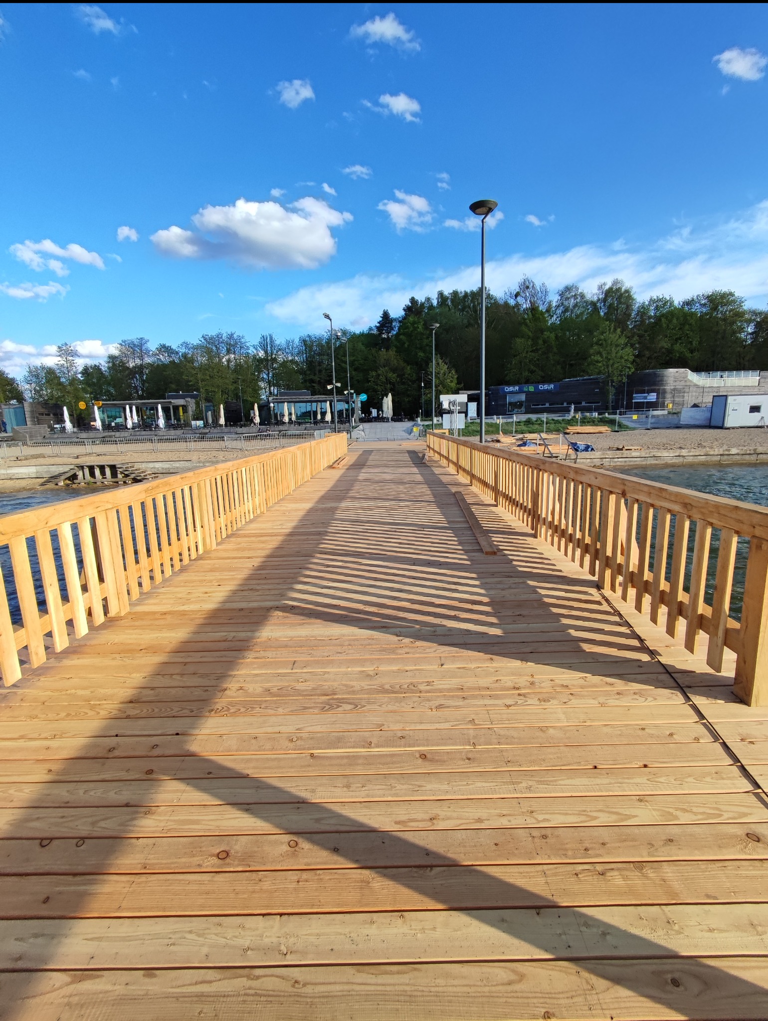 Nowy drewniany pomost z jasnego drewna prowadzący nad wodą, z widocznymi cieniami rzucanymi przez barierki, w tle budynki i błękitne niebo.