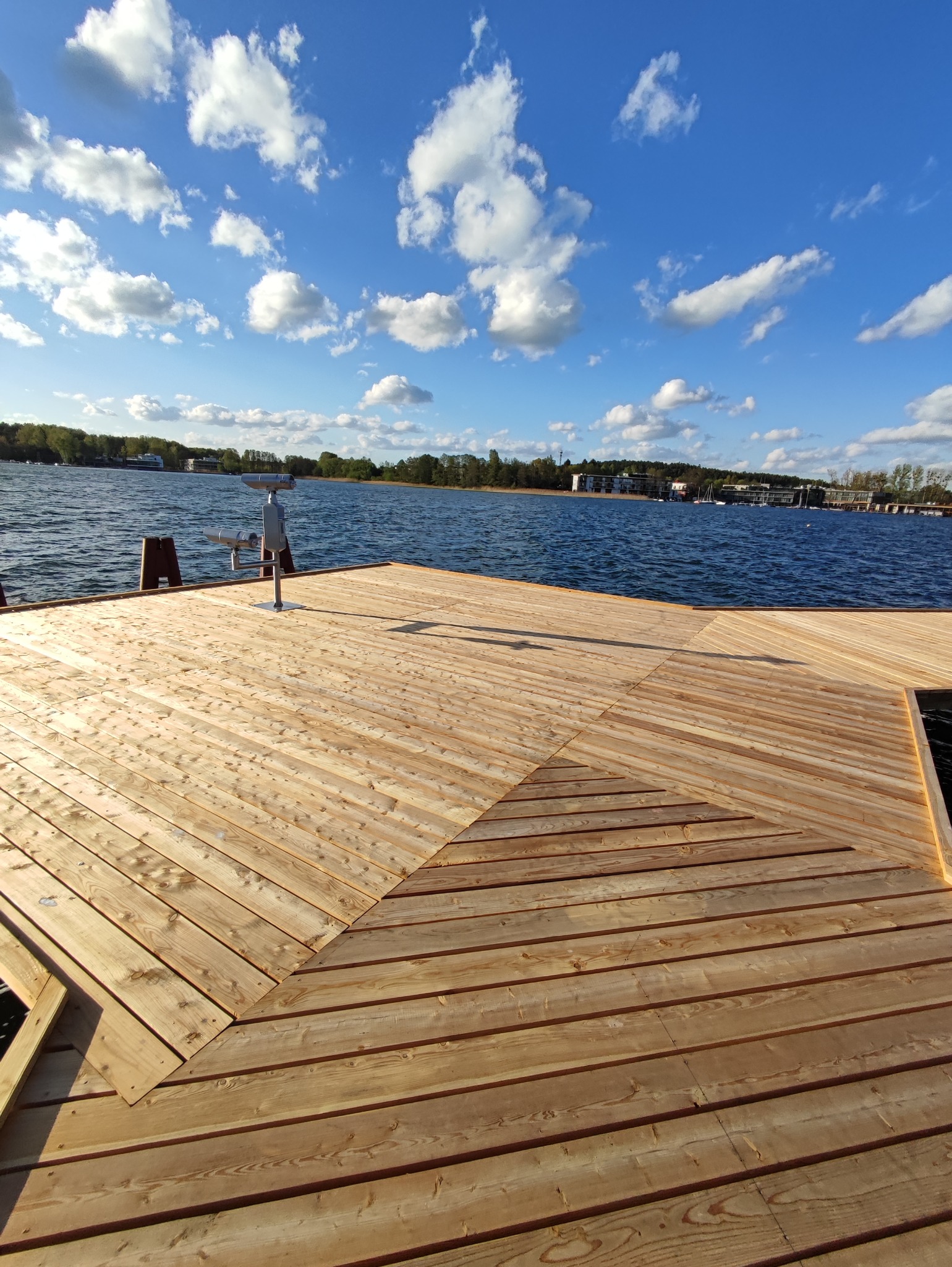 Jasny, drewniany taras nad jeziorem z metalowym teleskopem widokowym, błękitne niebo z chmurami w tle.