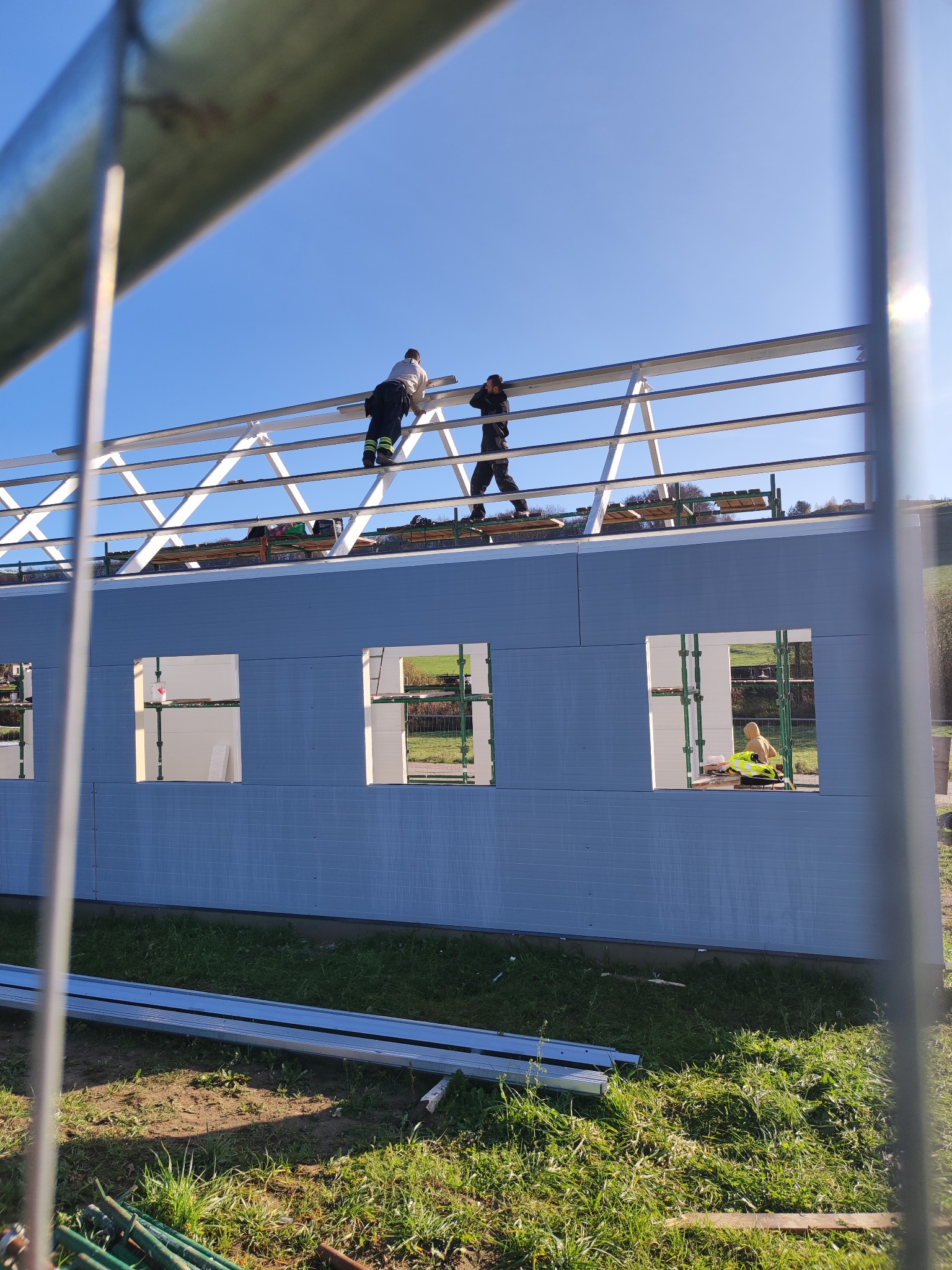 Montaż konstrukcji stalowej dachu na budynku w trakcie budowy, widoczne panele ścienne i pracownicy na górze.