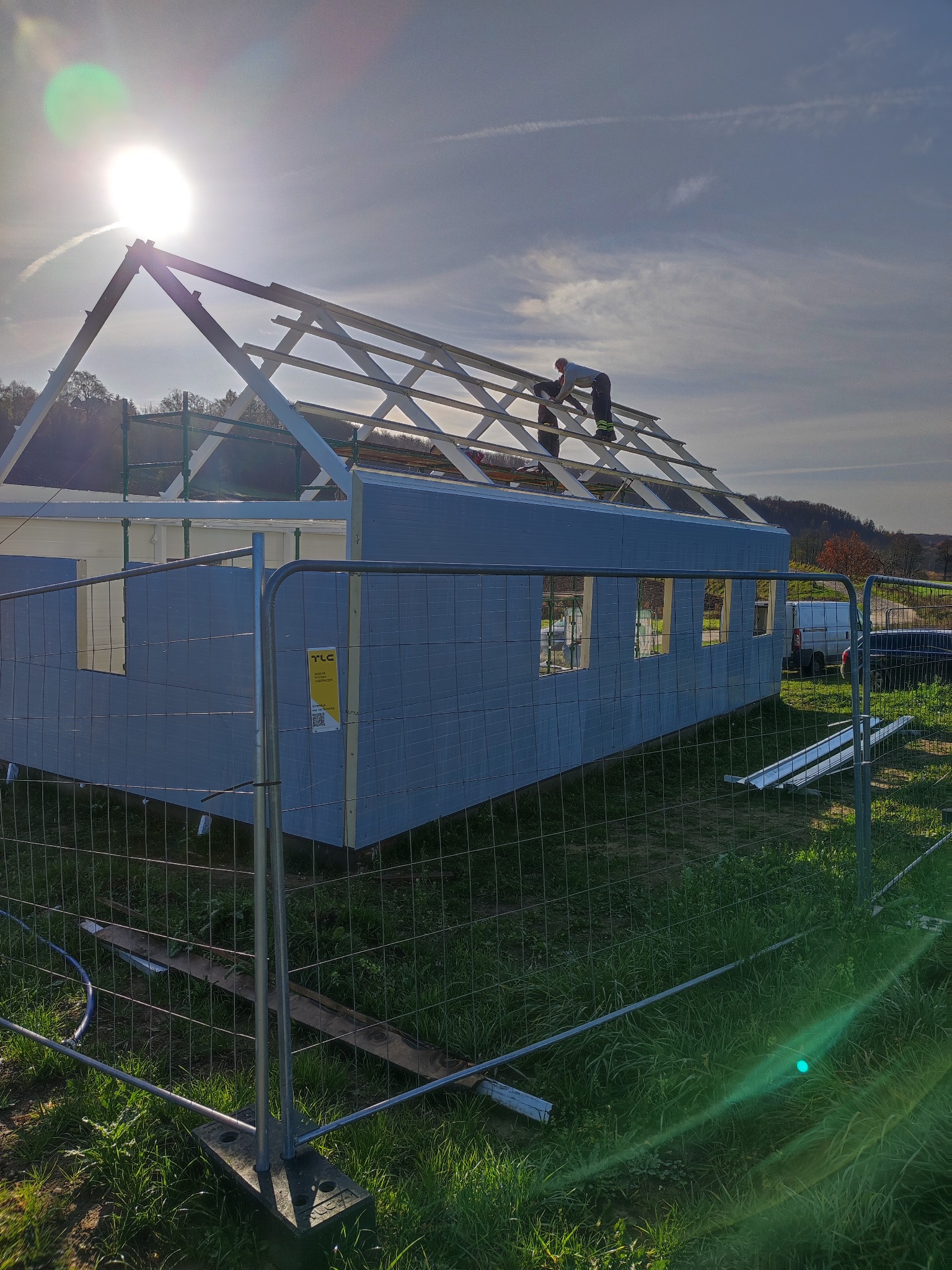 Wznoszenie szkieletu domu z prefabrykatów na tle zachodzącego słońca, widoczni dwaj pracownicy na dachu, ogrodzenie budowy z siatki.