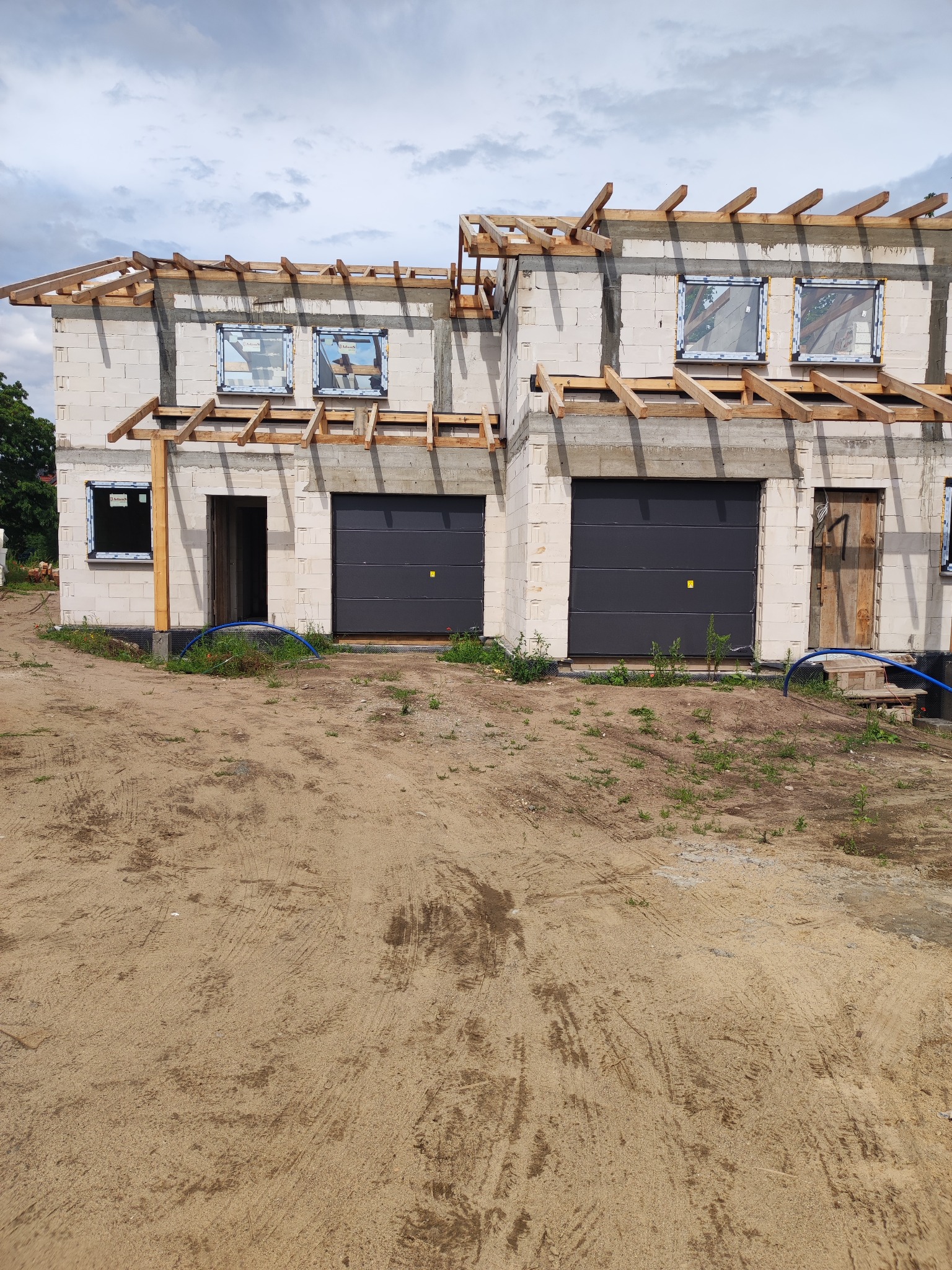 Surowy stan dwukondygnacyjnego domu z pustaków, z zamontowanymi oknami i drewnianą konstrukcją dachu, garaże z szarymi bramami, teren wokół w stanie naturalnym.