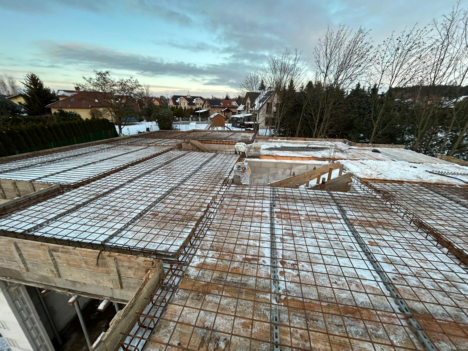 Zimowy widok na zbrojenie stropu domu w trakcie budowy, pokryte cienką warstwą śniegu, z widocznymi bloczkami betonowymi i szalunkiem drewnianym, w tle zabudowa jednorodzinna.