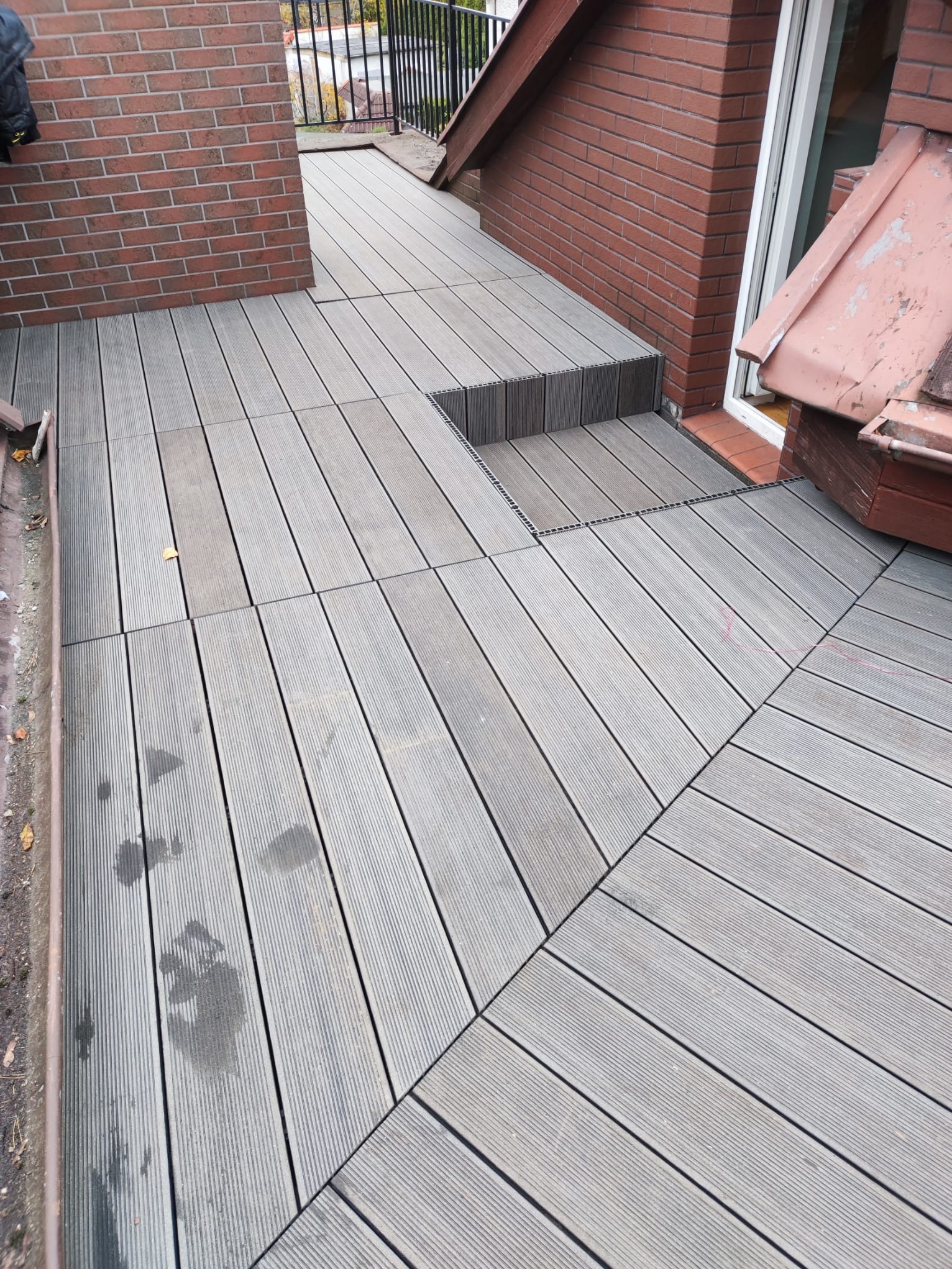Szary taras z desek kompozytowych, ułożonych w kwadraty, z widocznymi śladami wilgoci i pojedynczym liściem, przylegający do ceglanego budynku z oknem.