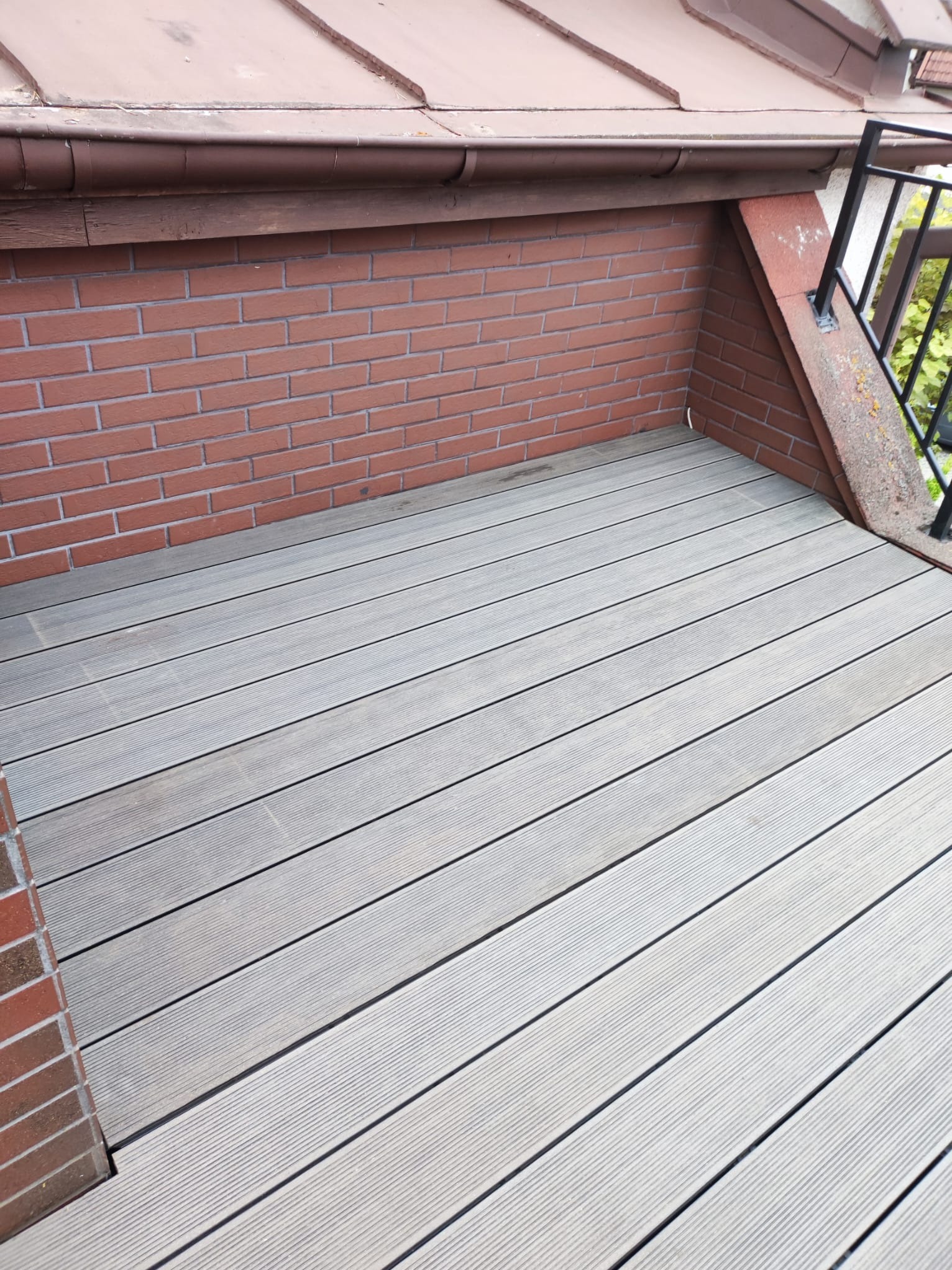 Szare deski tarasowe ułożone na balkonie z ceglaną ścianą i metalową barierką, widoczne z góry pod kątem.