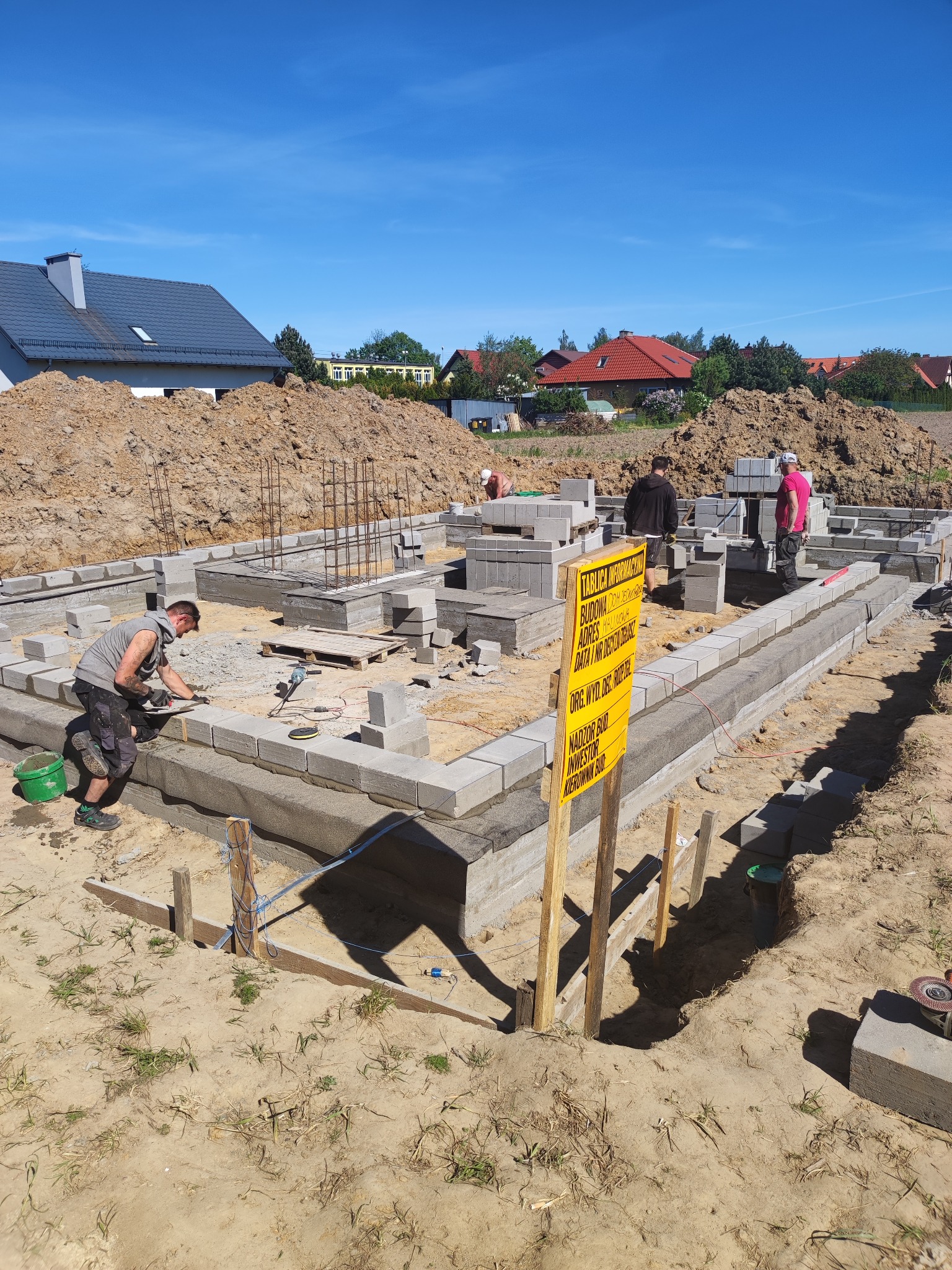 Budowa fundamentów domu z bloczków betonowych; widoczni pracownicy, tablica informacyjna budowy, zbrojenie pionowe, stos bloczków, narzędzia murarskie i hałda ziemi w tle.