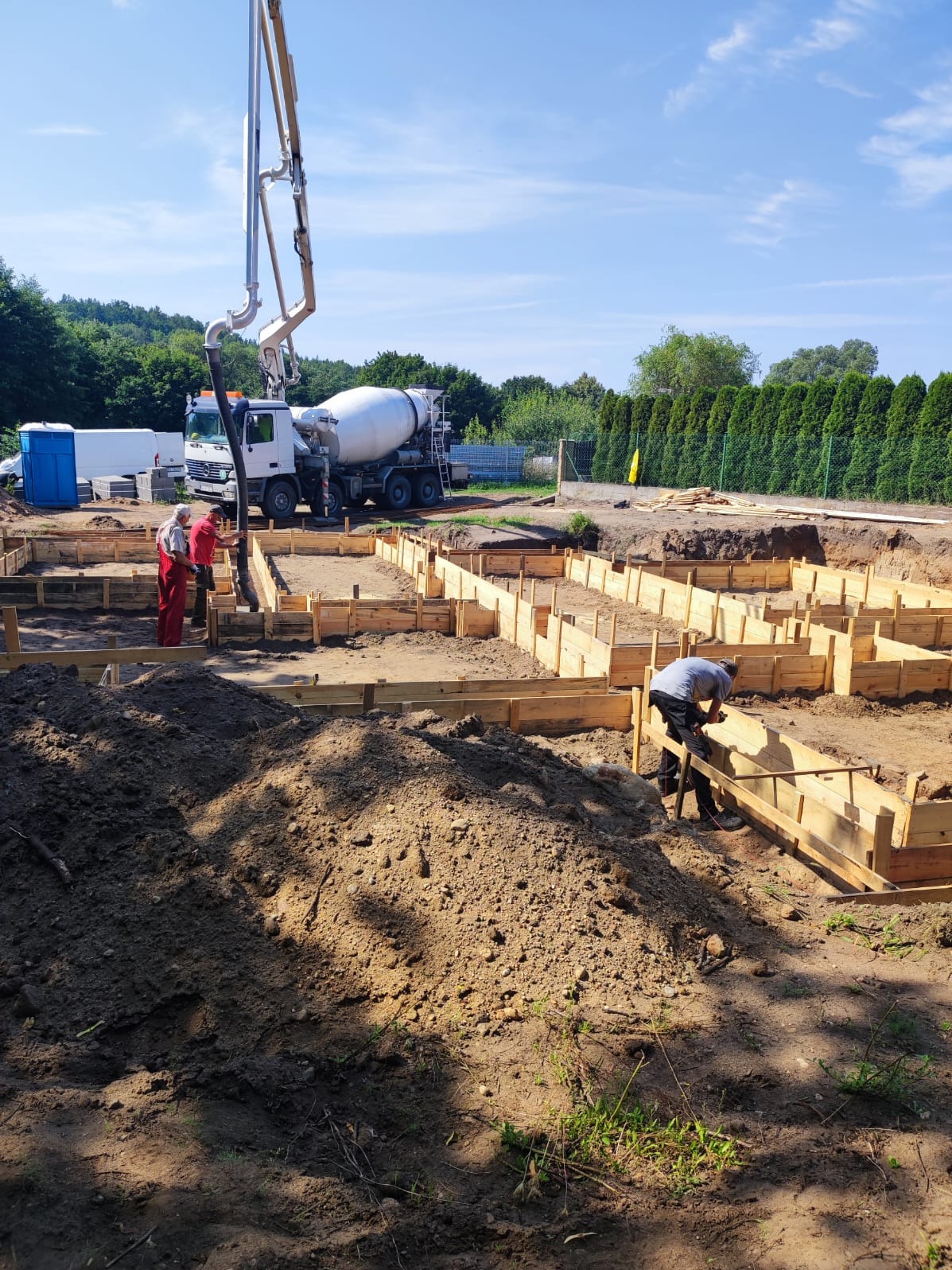 Wylewanie fundamentów pod budynek: beton z betoniarki pompowany do szalunków, pracownicy budowlani w kombinezonach, ziemia i wykop pod fundamenty.