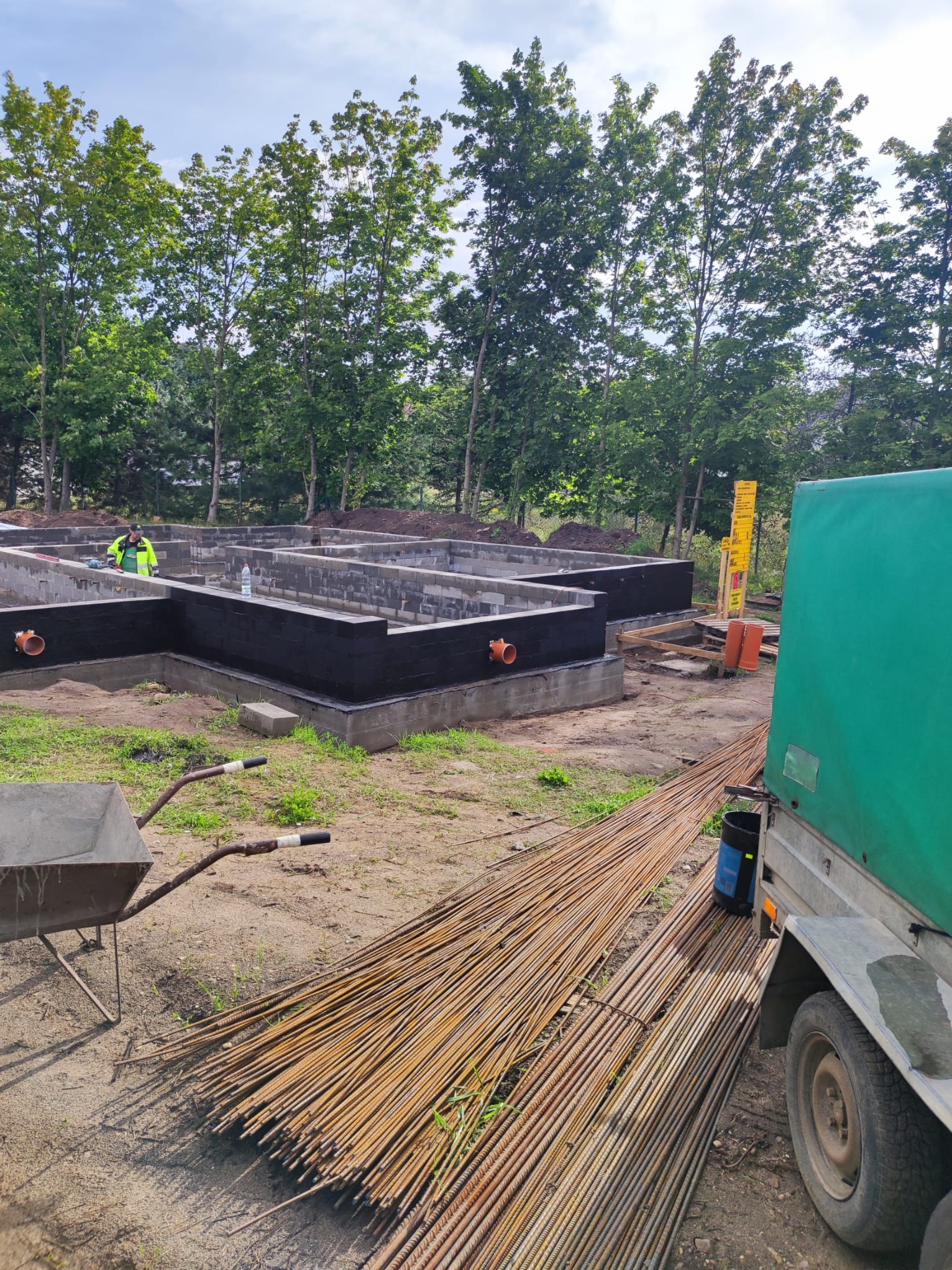 Fundamenty domu w trakcie budowy, z hydroizolacją, widoczna sterta prętów zbrojeniowych, taczka i przyczepka z zieloną plandeką w tle drzewa.