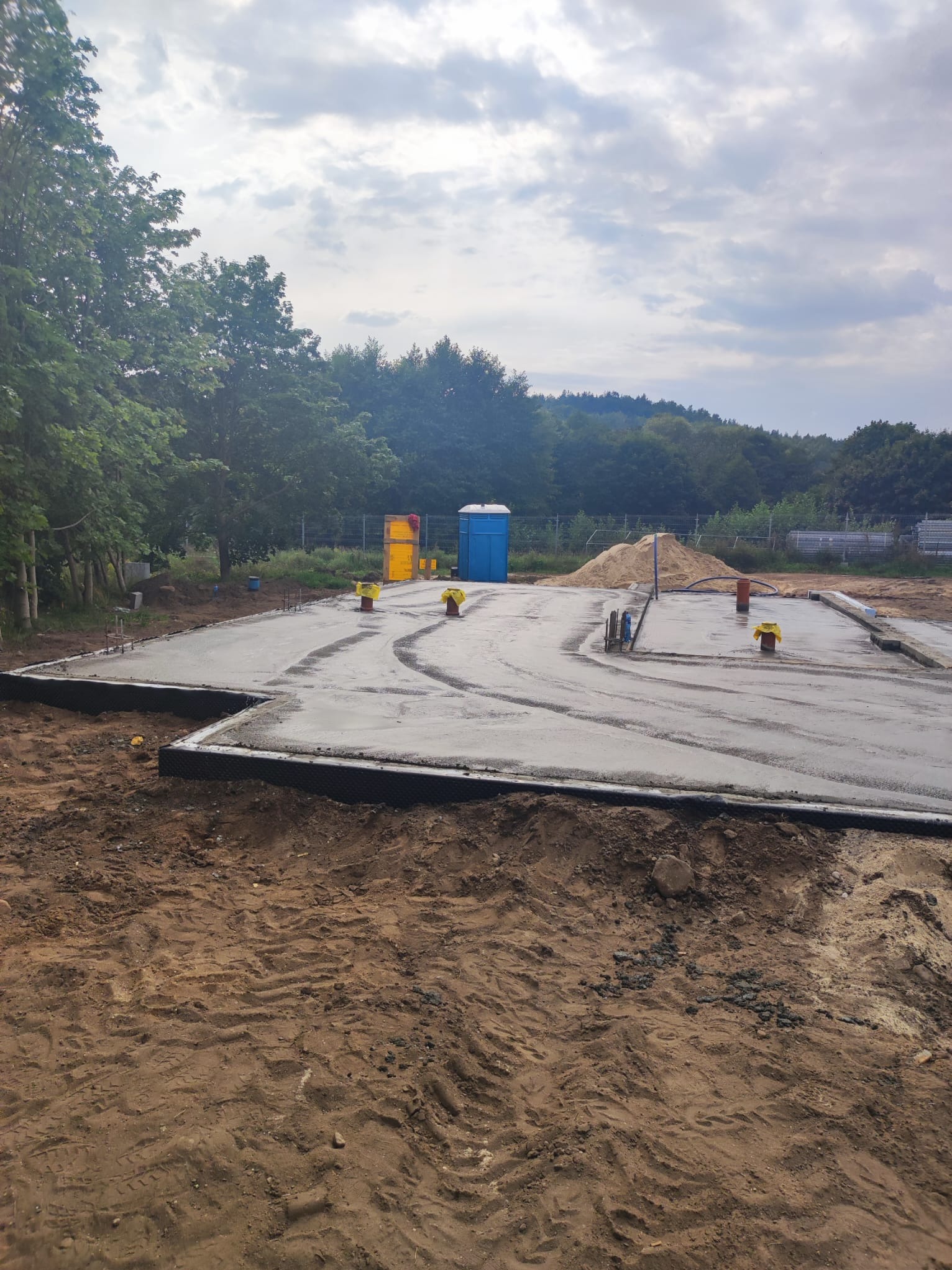 Wylana betonowa płyta fundamentowa z wystającymi rurami i zbrojeniami, widoczna izolacja pionowa, w tle przenośna toaleta i pryzma piasku na placu budowy.