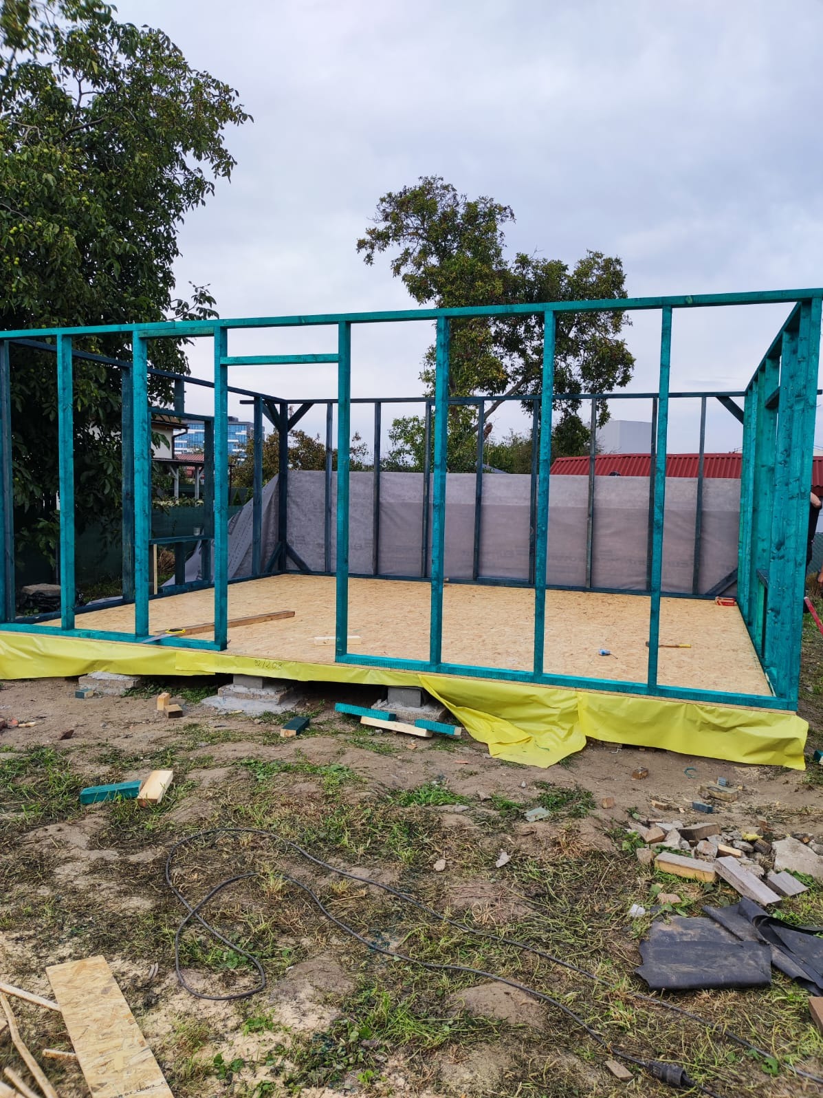 Wznoszenie szkieletu domu: widok na pomalowaną na turkusowo drewnianą konstrukcję ścian z płytami OSB na podłodze, zabezpieczoną żółtą folią budowlaną, na tle zieleni i pochmurnego nieba.
