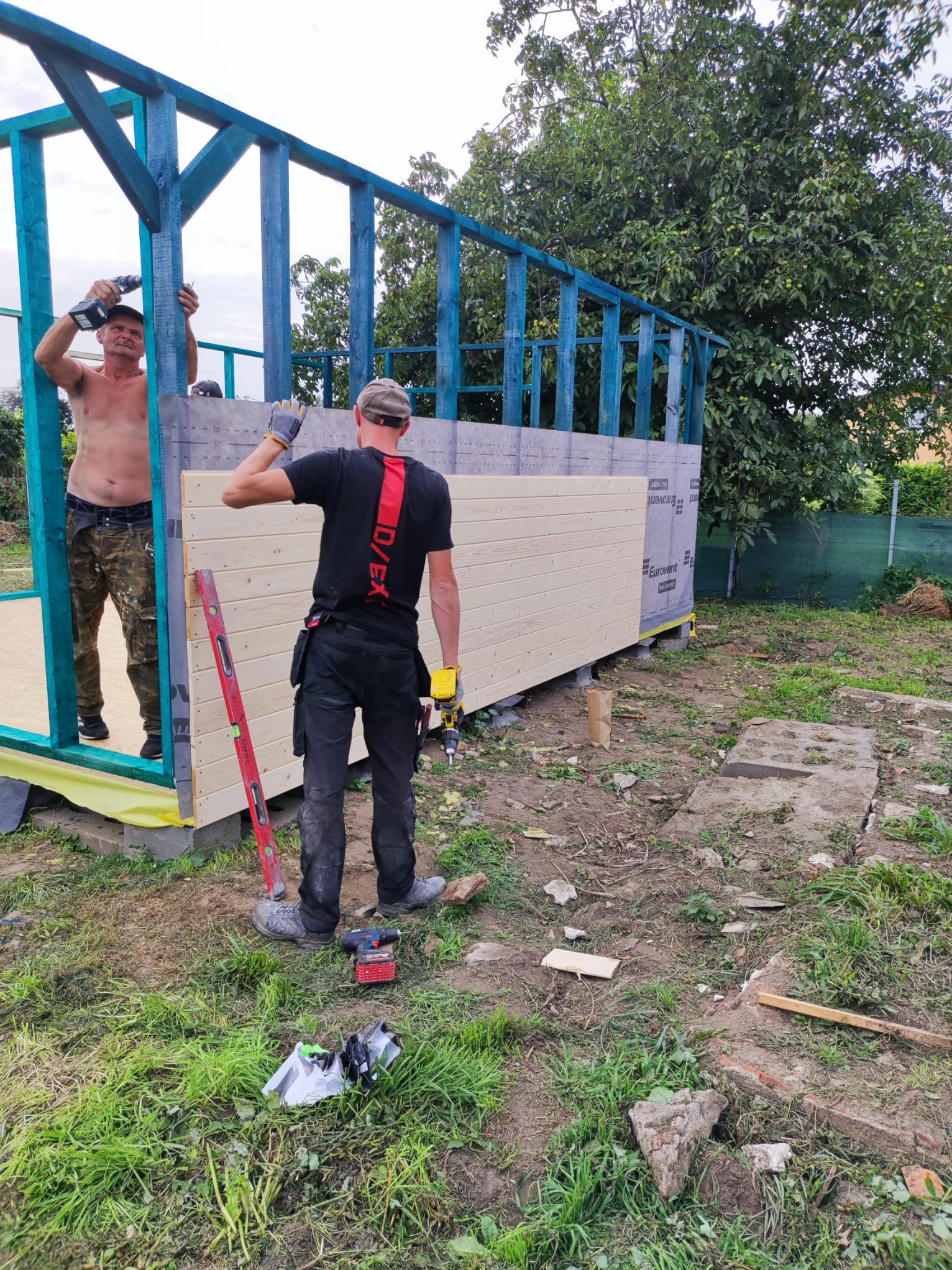 Dwóch mężczyzn montuje drewnianą konstrukcję szkieletową z niebieskimi elementami, jeden przykręca śruby od wewnątrz, drugi przykłada poziomicę od zewnątrz, widoczne narzędzia i materiały budowlane.