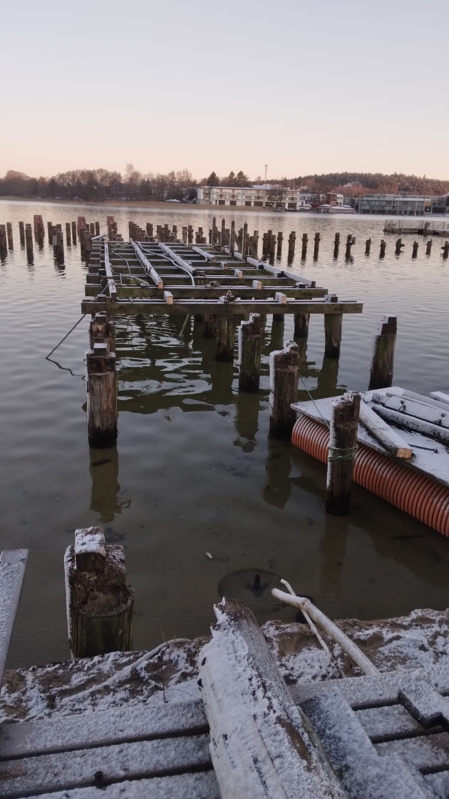 Zimowy widok na częściowo zniszczony drewniany pomost na jeziorze, pokryty świeżym śniegiem, z widocznymi fragmentami konstrukcji i palikami wystającymi z wody, na tle spokojnej tafli jeziora...