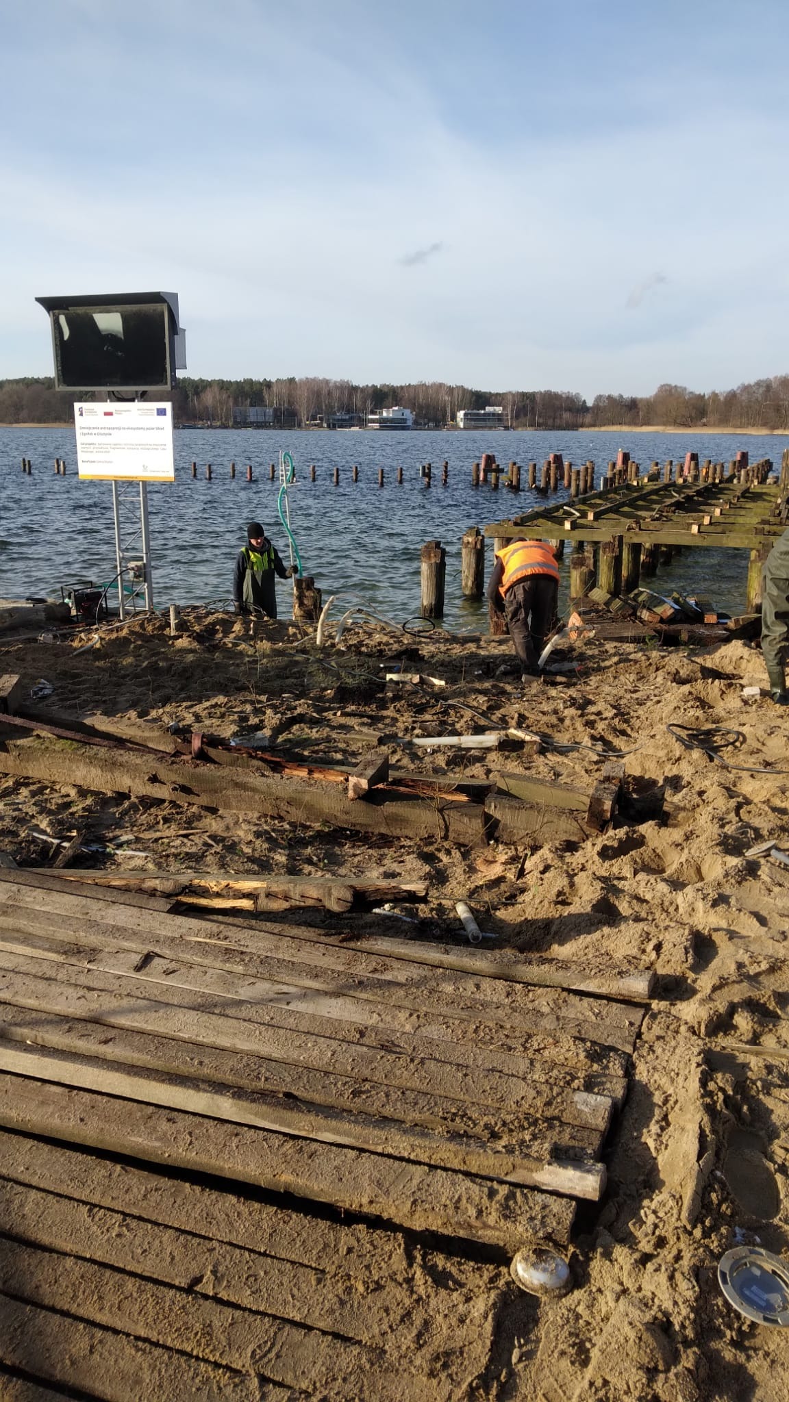 Demontaż starego drewnianego pomostu nad jeziorem, pracownicy w kamizelkach odblaskowych podczas pracy, widoczne resztki desek i konstrukcji, w tle woda i drzewa.